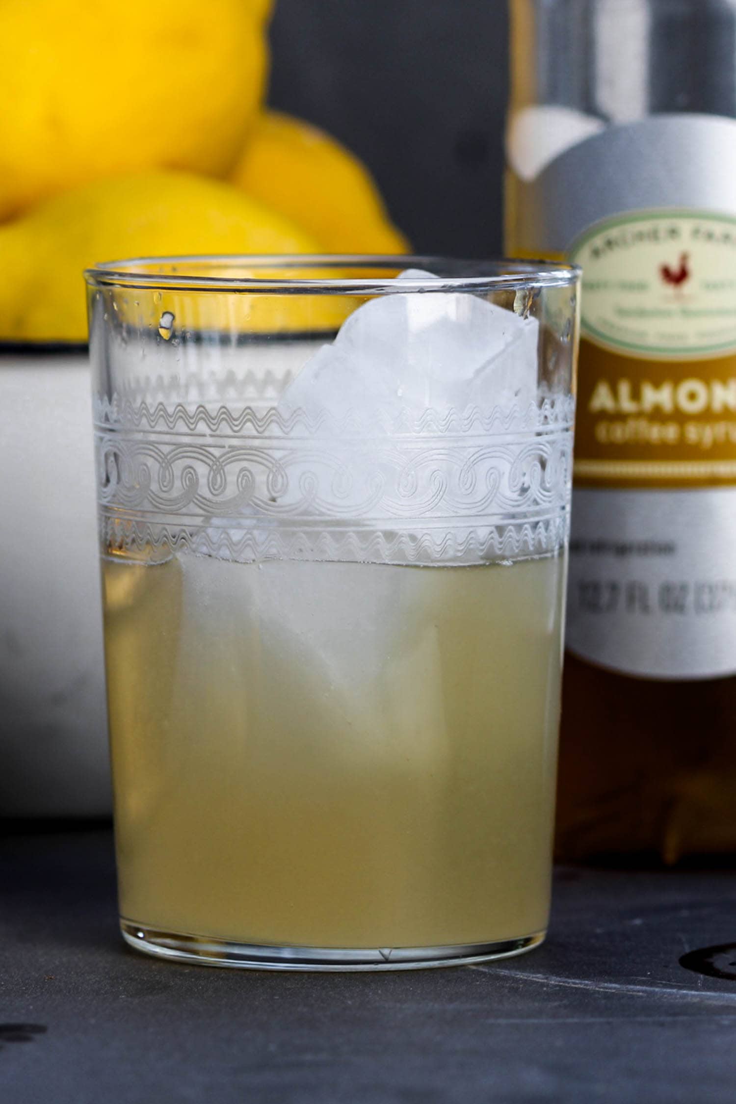 Almond syrup filling half of decorative glass filled with ice with bowl of lemons and bottle of almond syrup in background on a gray slate surface