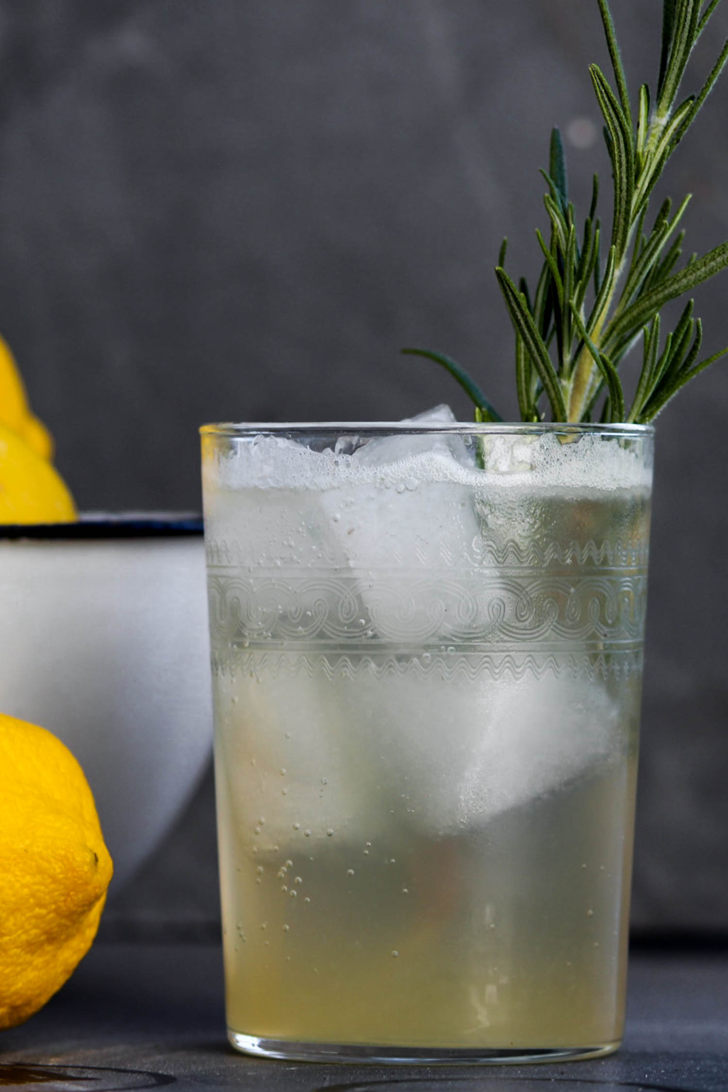 Glass of Venetian lemonade with ice and a sprig of rosemary sitting on blue slate surface beside of bowl of lemons