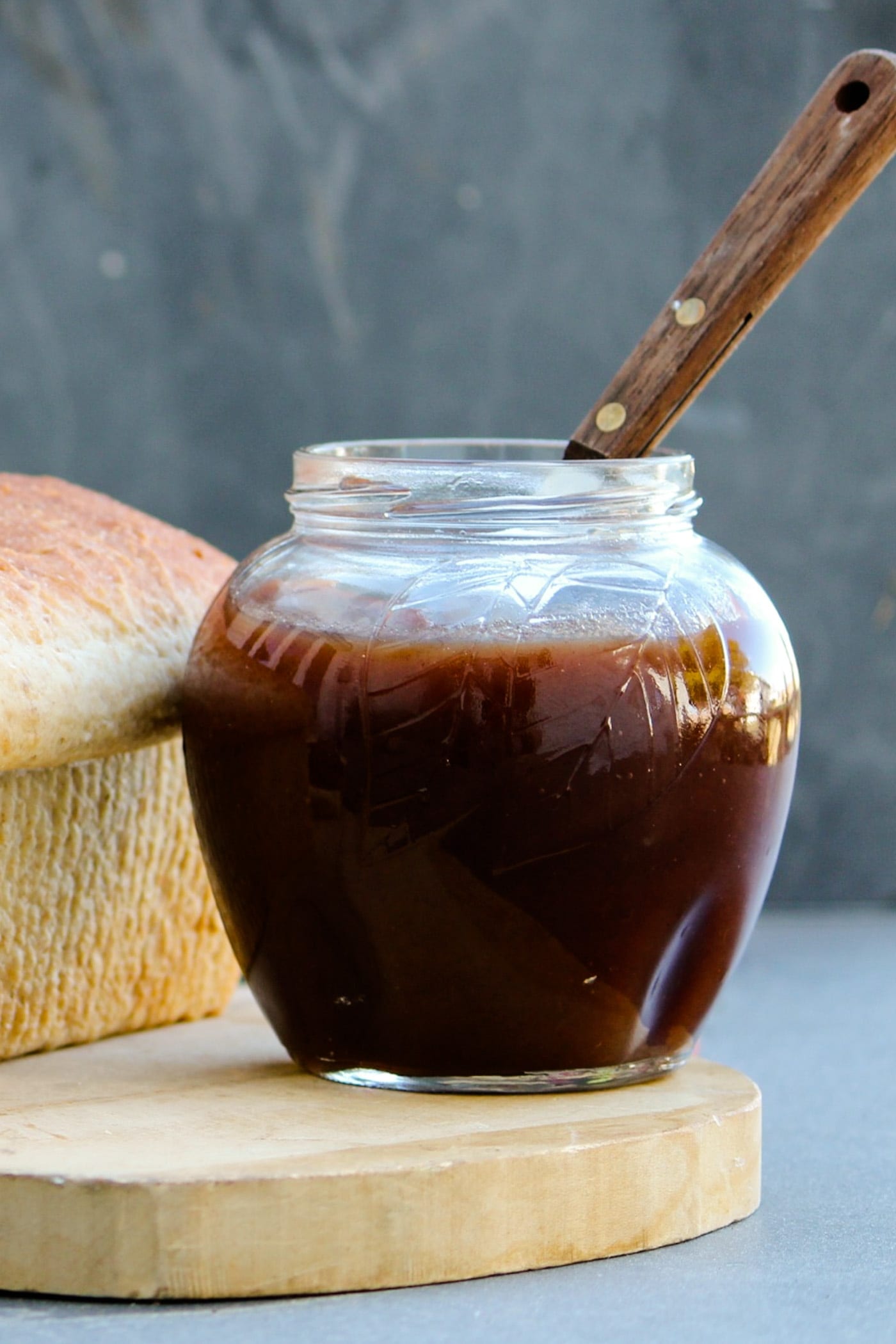 Jar of slow cooker apple butter with loaf of bread beside it and knife sitting on a wood cutting board in front of a gray slate surface