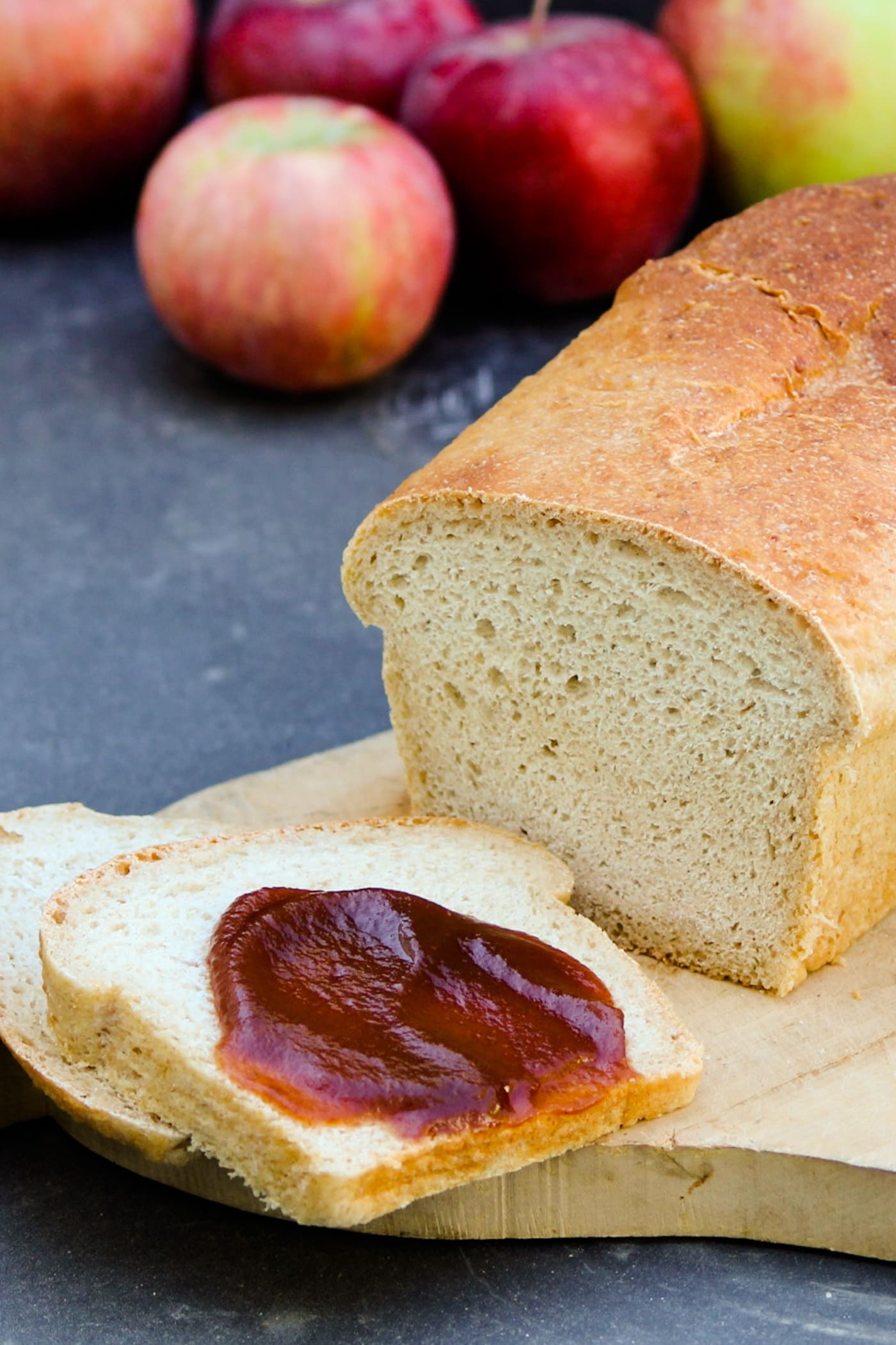 Slow cooker apple butter spread on piece of homemade white bread sitting on a cutting board with extra bread loaf behind on a gray slate surface with extra apples in background