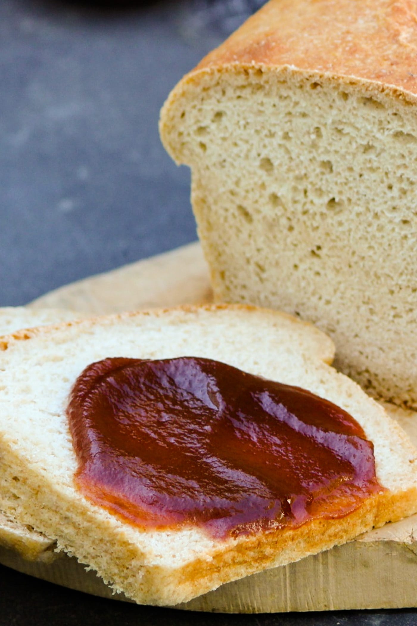Slow cooker apple butter spread on piece of homemade white bread sitting on a cutting board with extra bread loaf behind on a gray slate surface