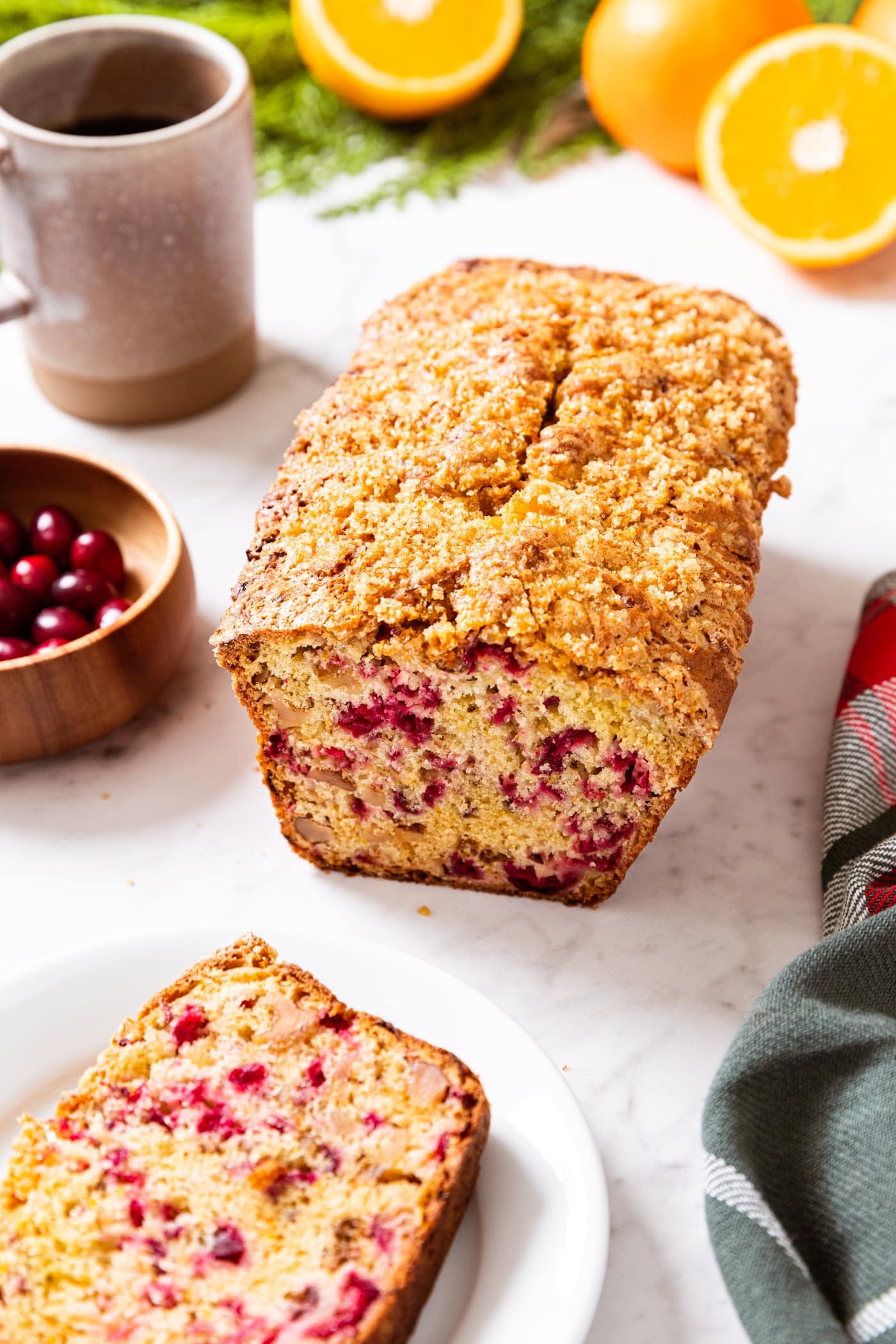 Loaf of cranberry orange bread sitting on white marble surface with slice of bread sitting on white plate with cup of coffee in the background along with oranges and cranberries.