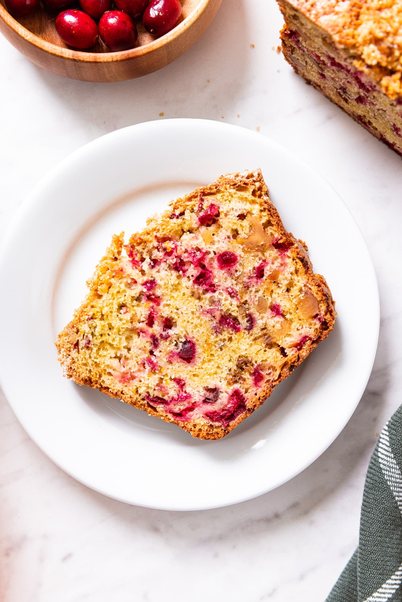 White marble surface with plate sitting with slice of cranberry orange bread with cranberries and rest of loaf of bread to the side.