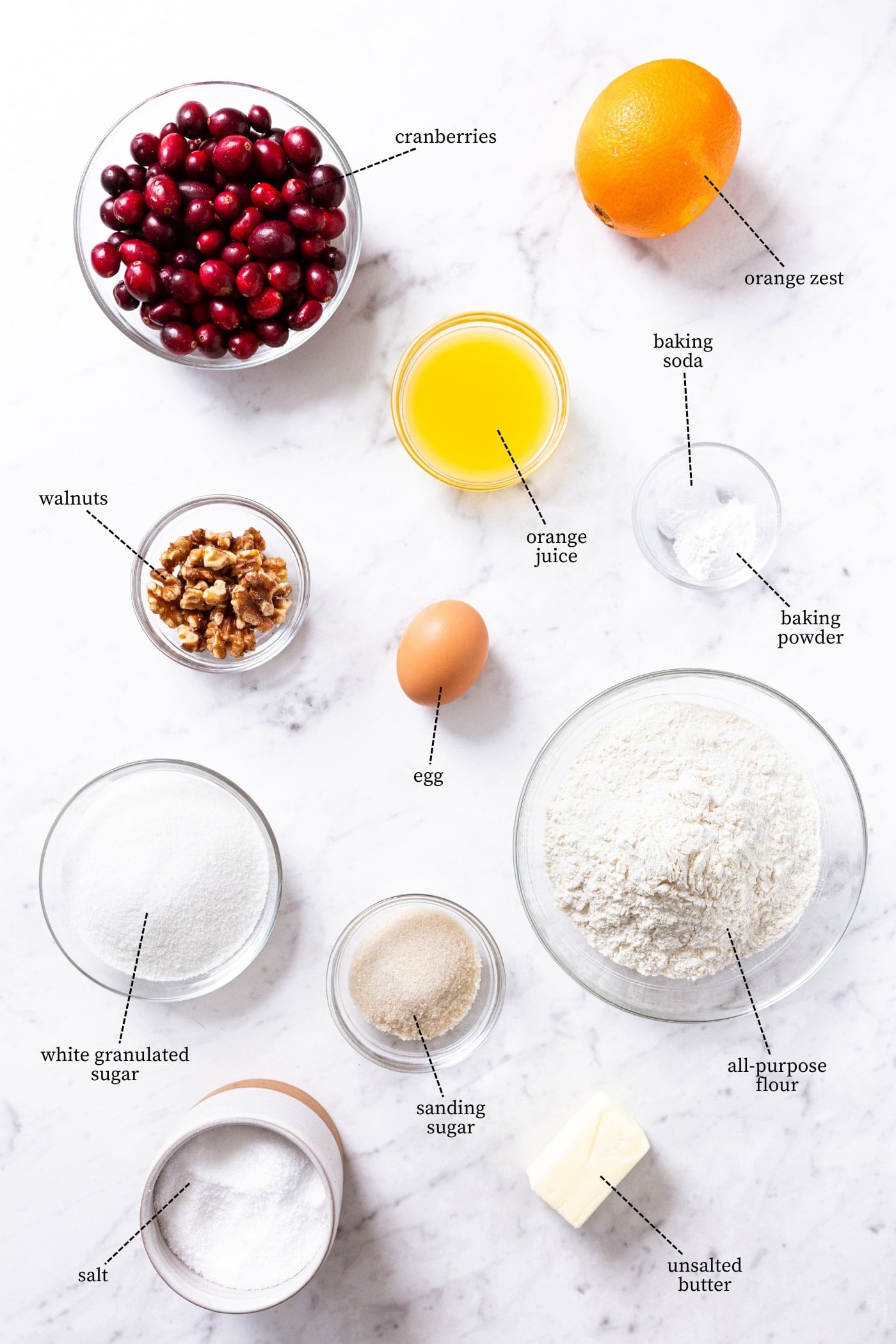 White marble surface filled with all ingredients needed to make cranberry orange bread including cranberries, orange juice, orange, flour, sugar, spices, and more.