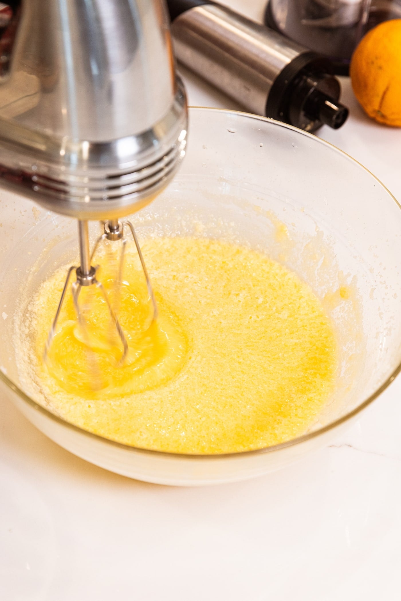 Hand mixer mixing together batter with light orange color on white surface with orange in the background.