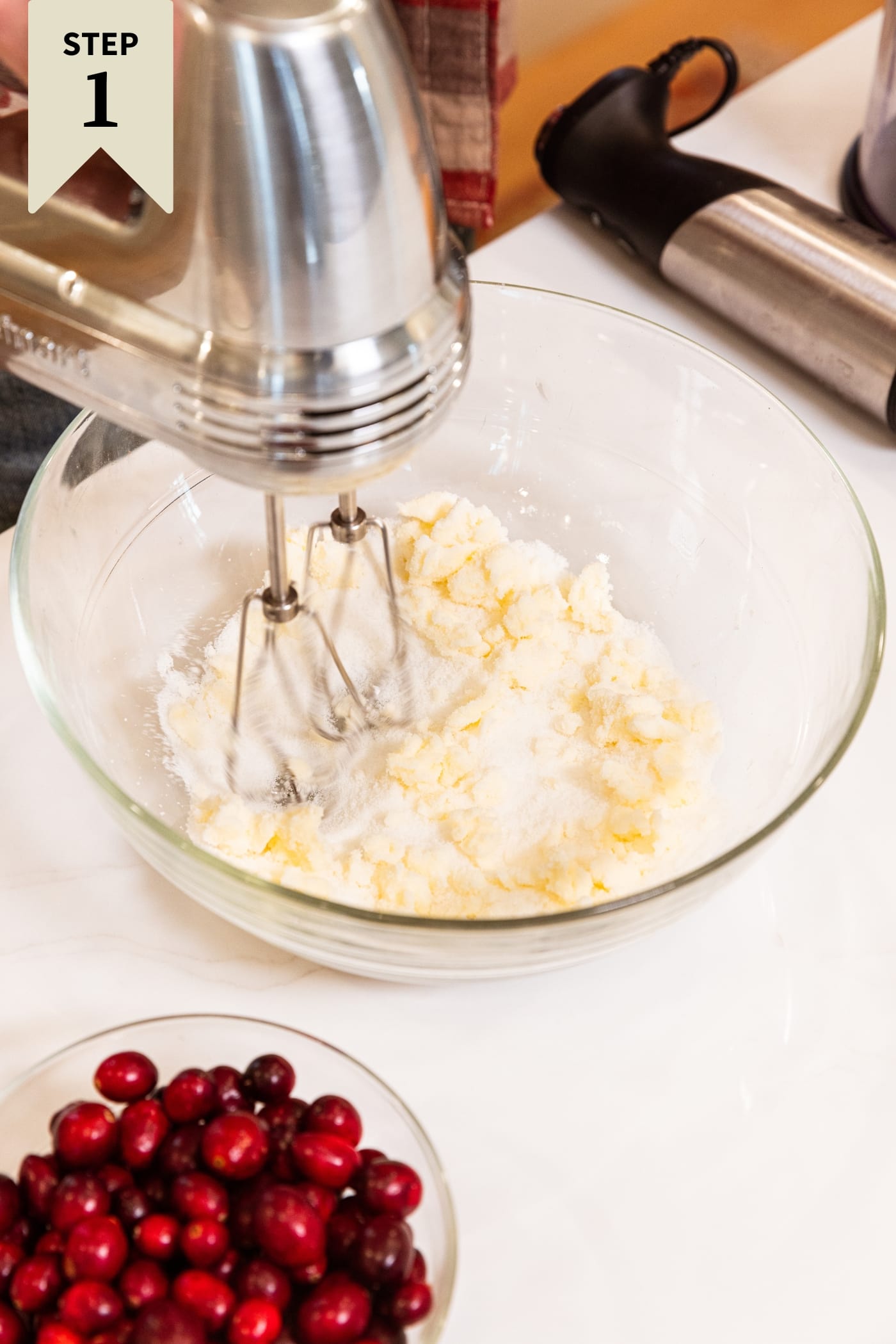 Glass mixing bowl with hand mixer spinning inside with butter and sugar being mixed together all on white surface with bowl of cranberries to the side.
