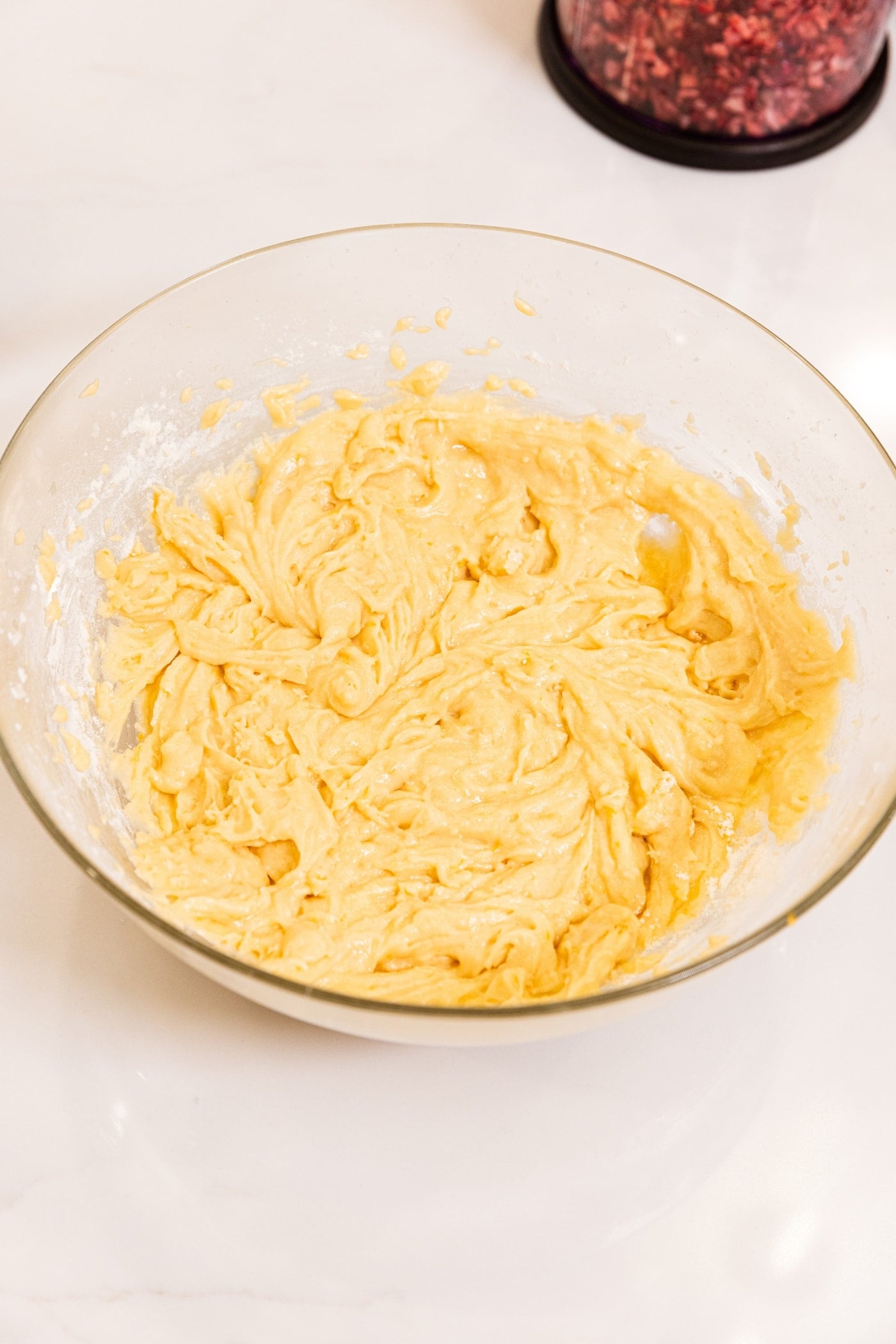 Glass bowl filled with light yellow colored batter after being mixed with a hand mixer sitting on white surface.