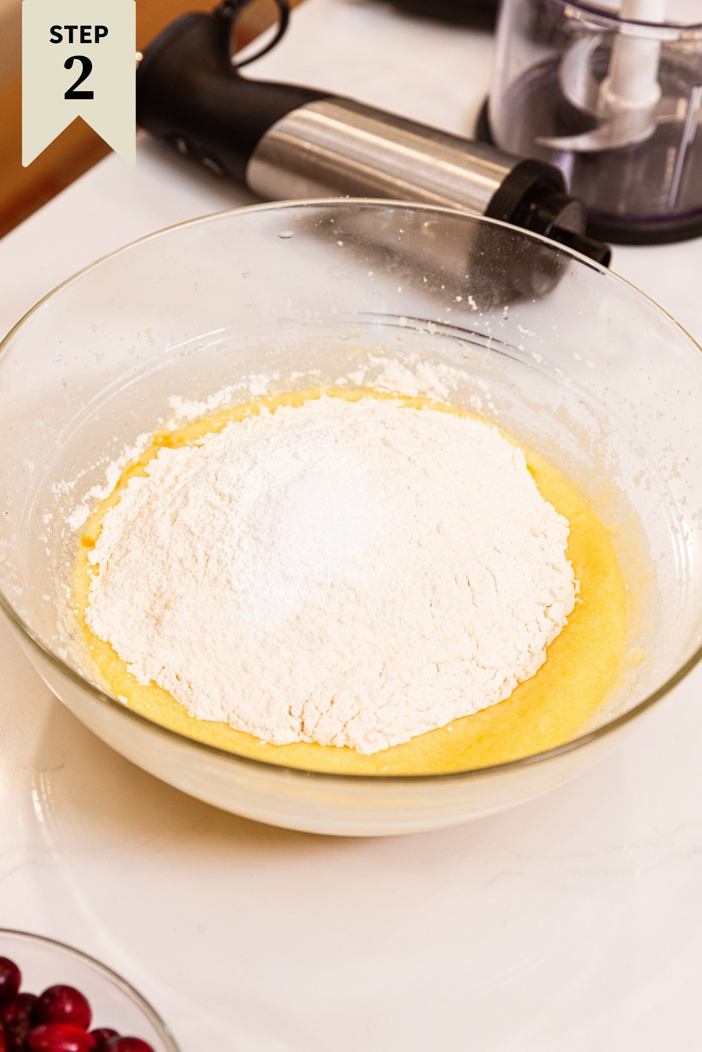 Glass mixing bowl filled with yellow colored batter with scoops of flour on top before being mixed with hand mixer all on white surface.