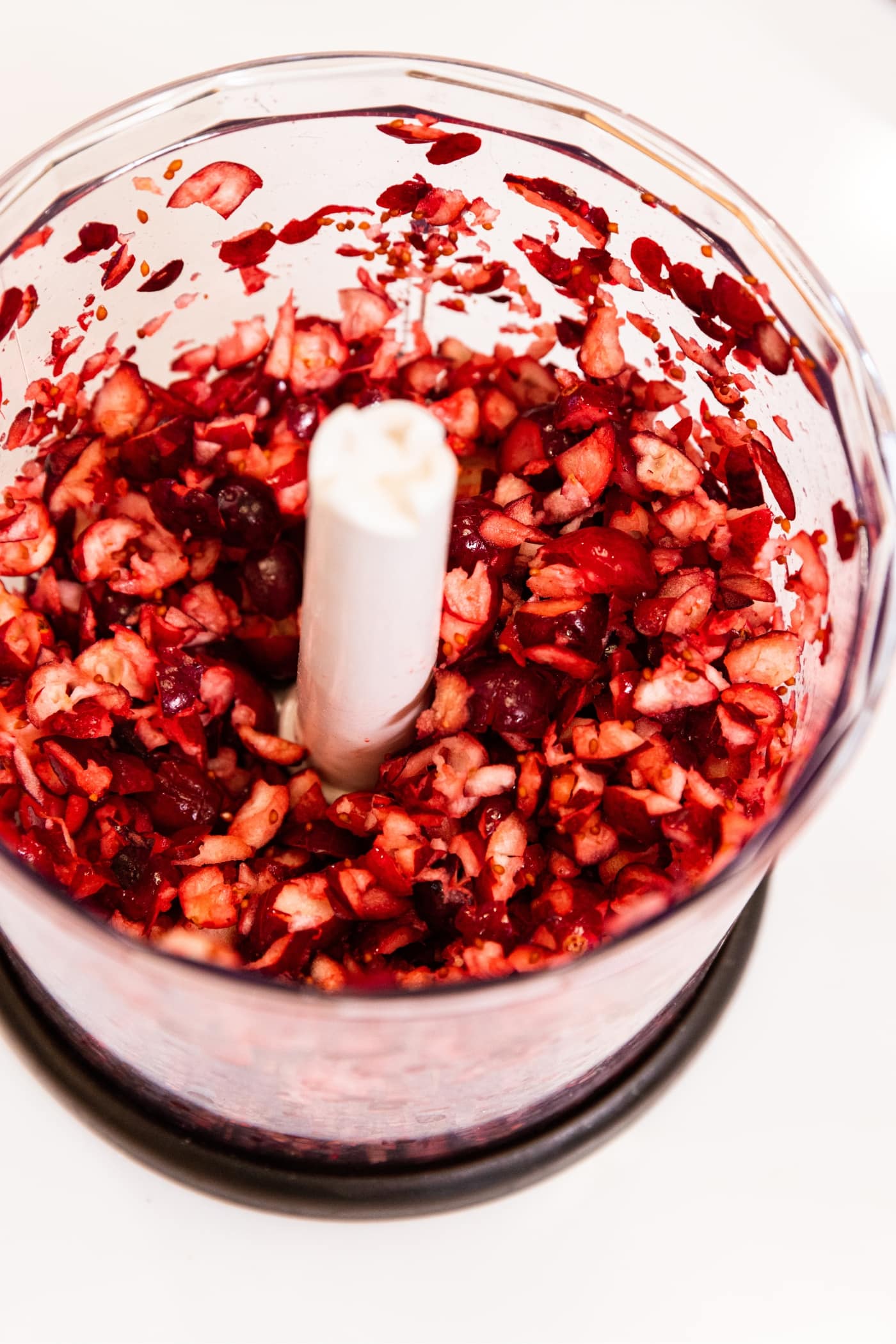 Small food processor filled with pieces of chopped fresh cranberries sitting on white surface.