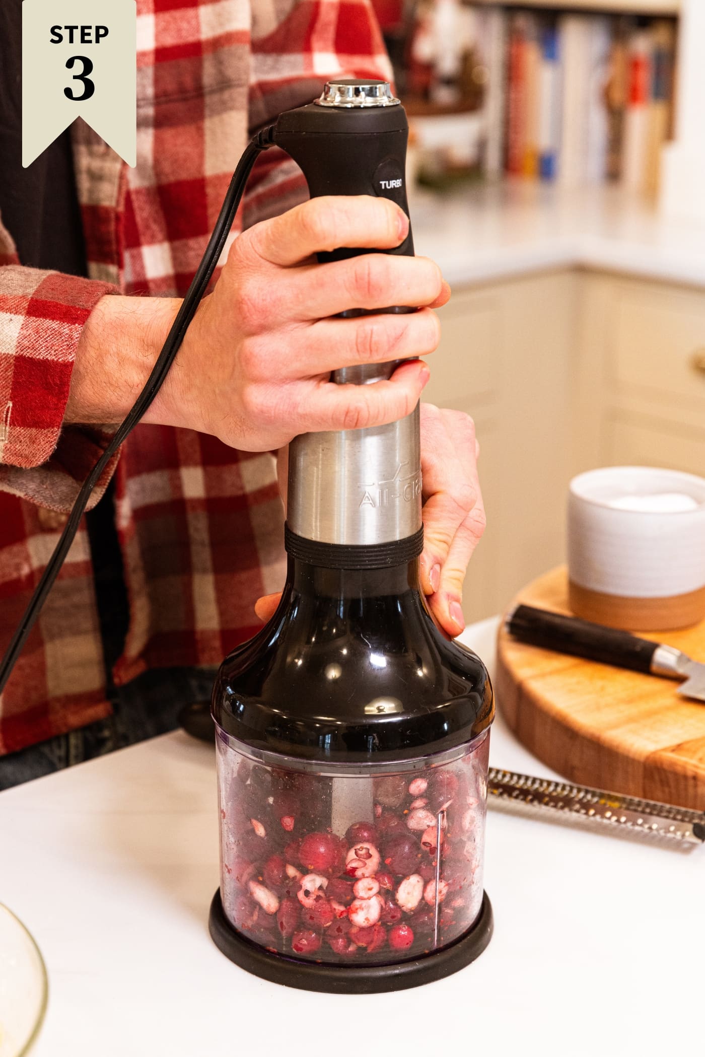 Hand holding food processor chopping up cranberries all on white surface with wood cutting board in background.