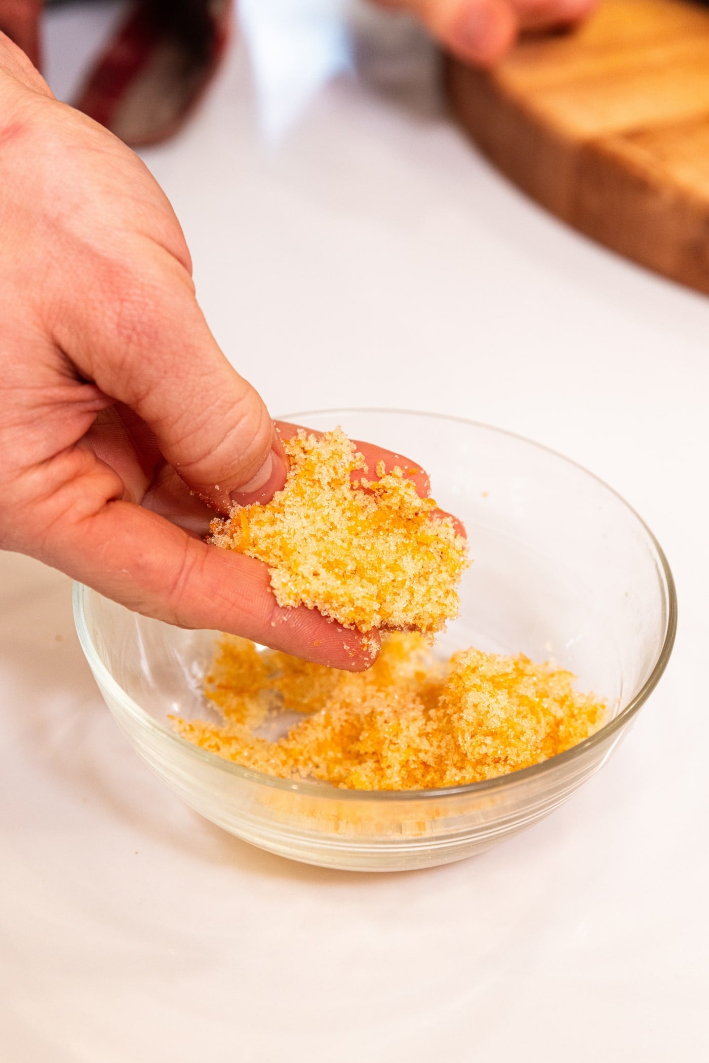 Hand holding orange zest and sanding sugar mixed together in small glass bowl sitting on white countertop.