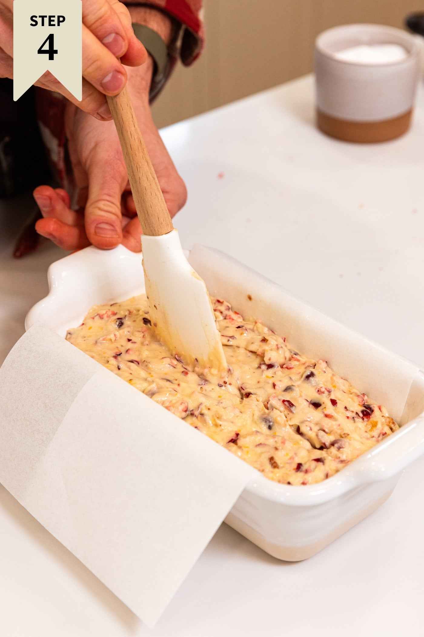 Hand pulling white spatula through the middle of cranberry orange bread dough in white loaf pan fitted with pieces of baking parchment all on white countertop.