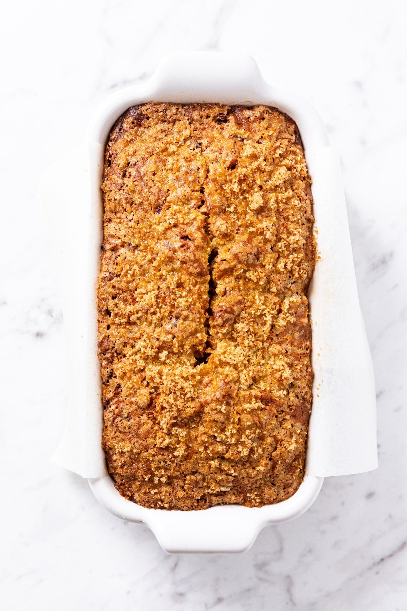 Baked orange cranberry bread sitting on white loaf pan on marble surface right after coming out of the oven.