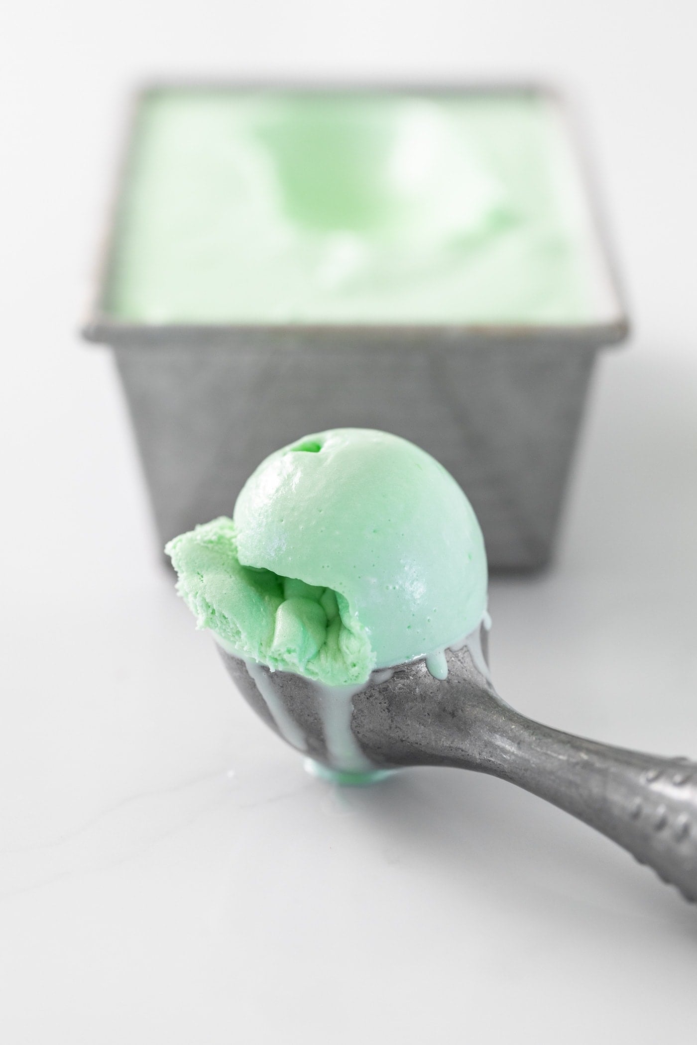 Gray ice cream scooper sitting on white countertop with green scoop of ice cream with gray pan in background