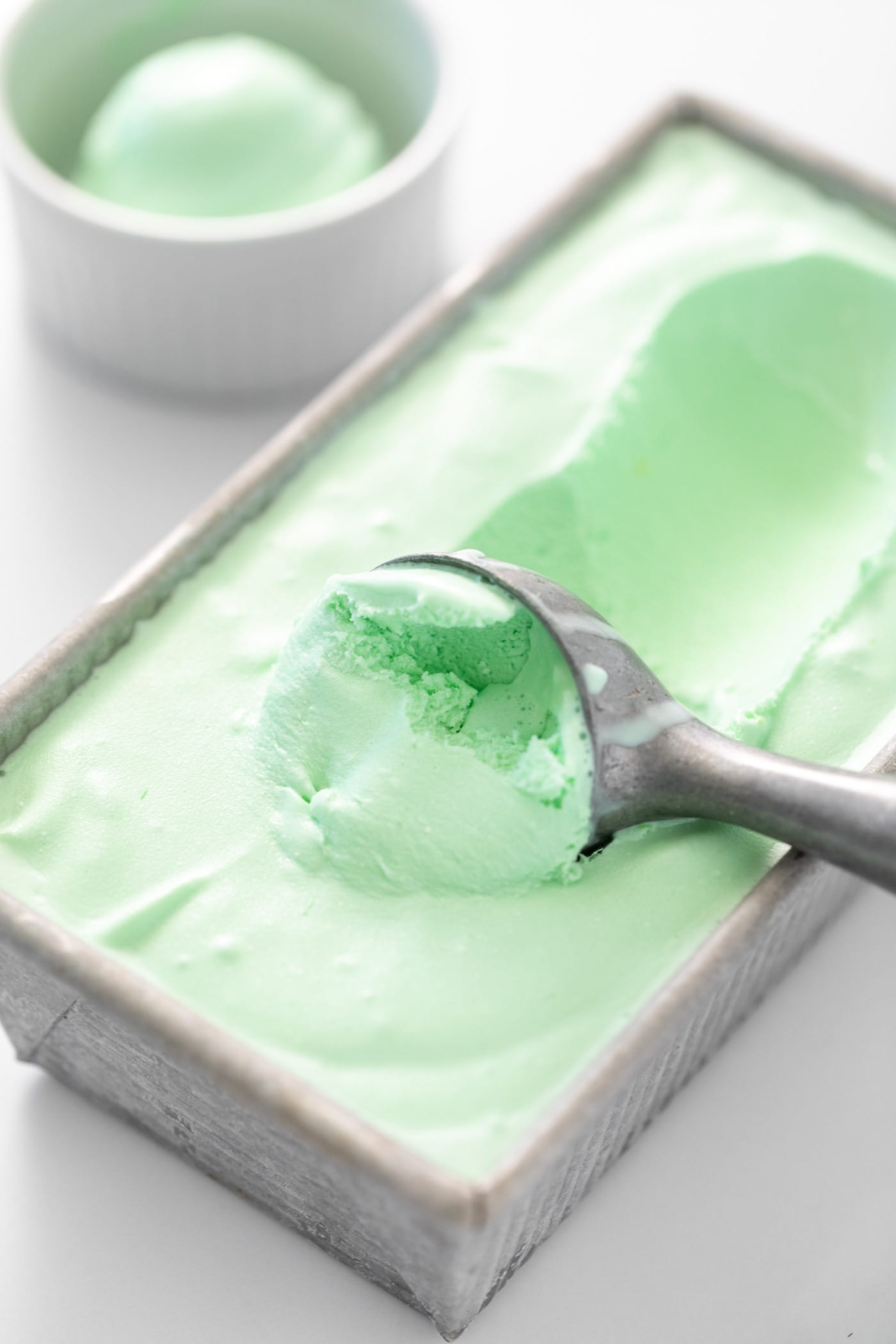 Ice cream scooper sitting in green colored ice cream in gray pan with small white bowl in background all on white surface