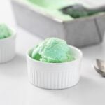 Green colored ice cream sitting in small white bowl with gray pan holding additional ice cream in background all on white surface