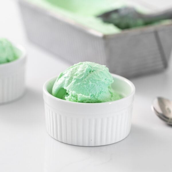 Green colored ice cream sitting in small white bowl with gray pan holding additional ice cream in background all on white surface
