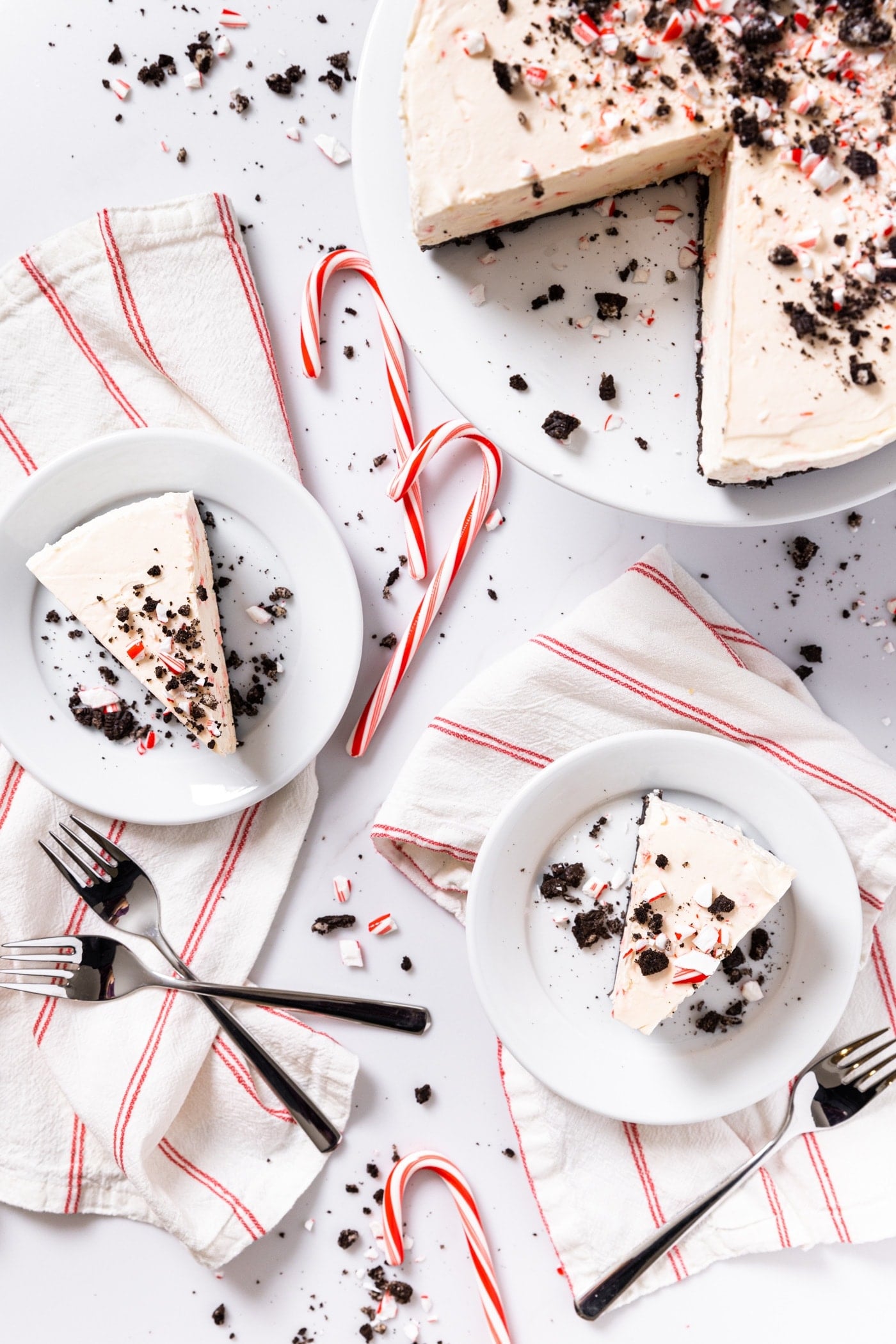 Top down view of slices of peppermint cheesecake sitting on white plates on white countertop filled with chocolate cookie crumbs and peppermint candy canes