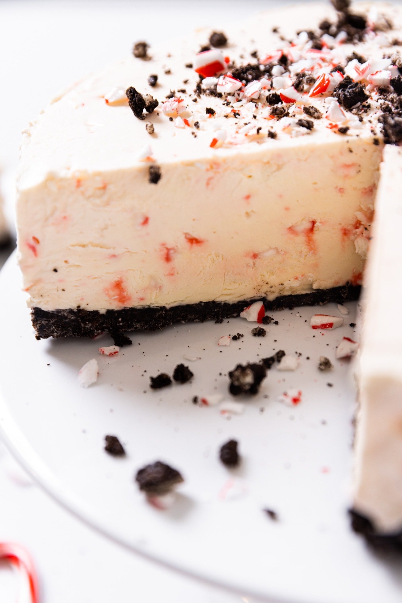 Close up view of peppermint cheesecake with slices taken out sitting on white cake plate with chocolate crumbles and candy cane pieces