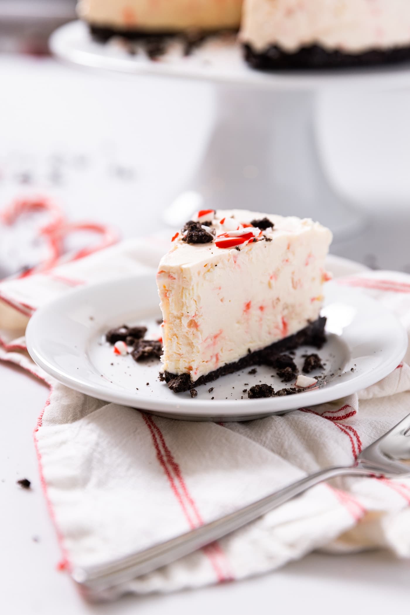 White plate with slice of peppermint cheesecake sitting on red colored napkin sitting on white countertop with cake plate in background