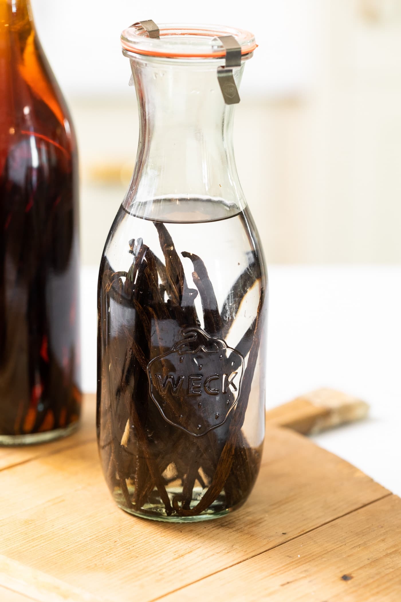 Large narrow bottle filled with clear alcohol sitting on wood board with vanilla beans inside of bottle