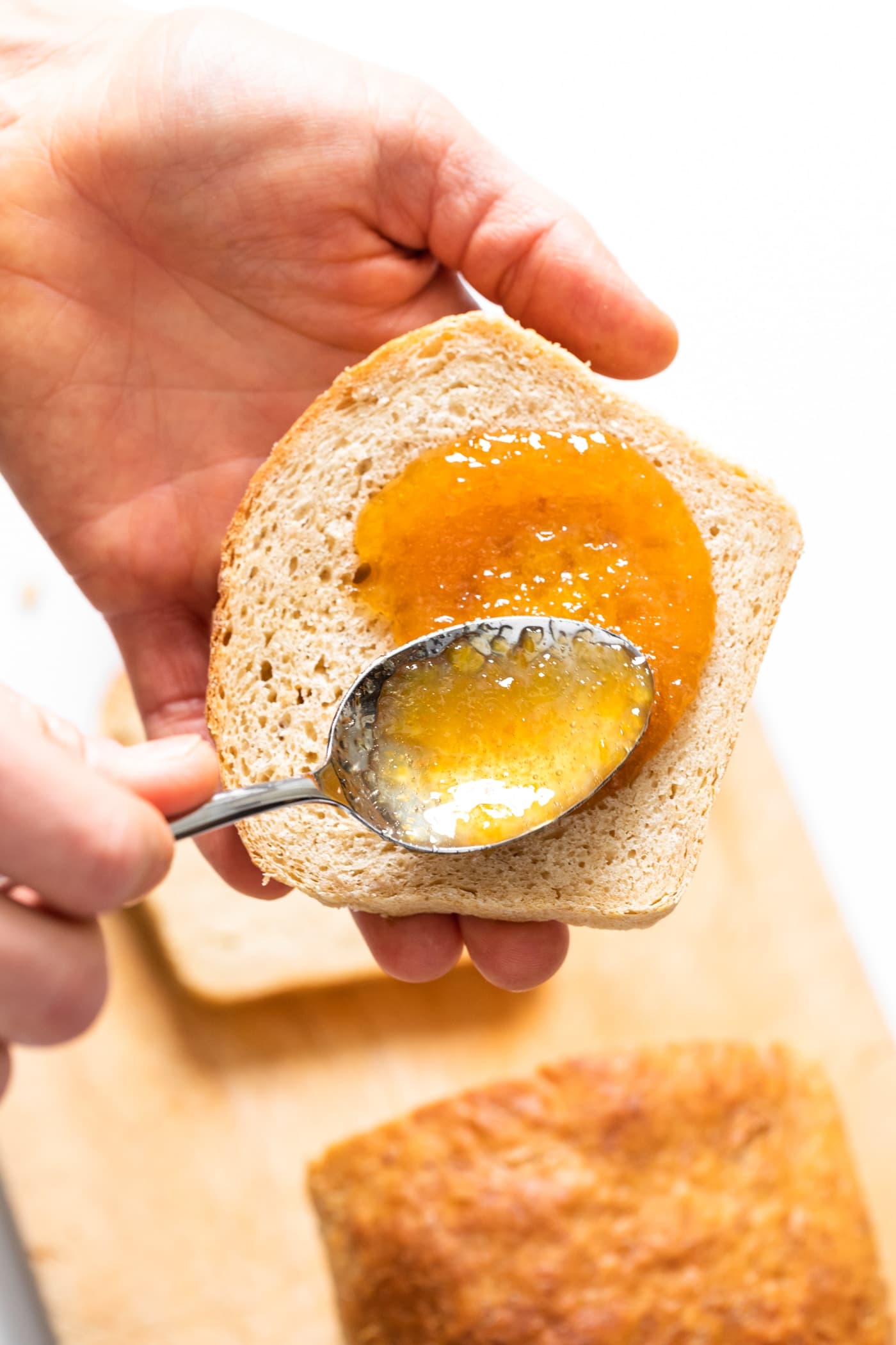 Slice of bread being slathered with orange colored jam with rest of loaf sitting in background