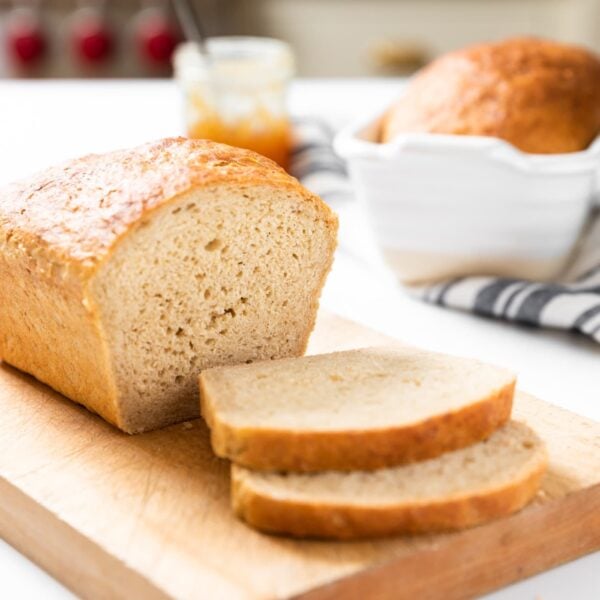 Loaf of bread with slices sitting on cutting board with jam jar in background all on white countertop