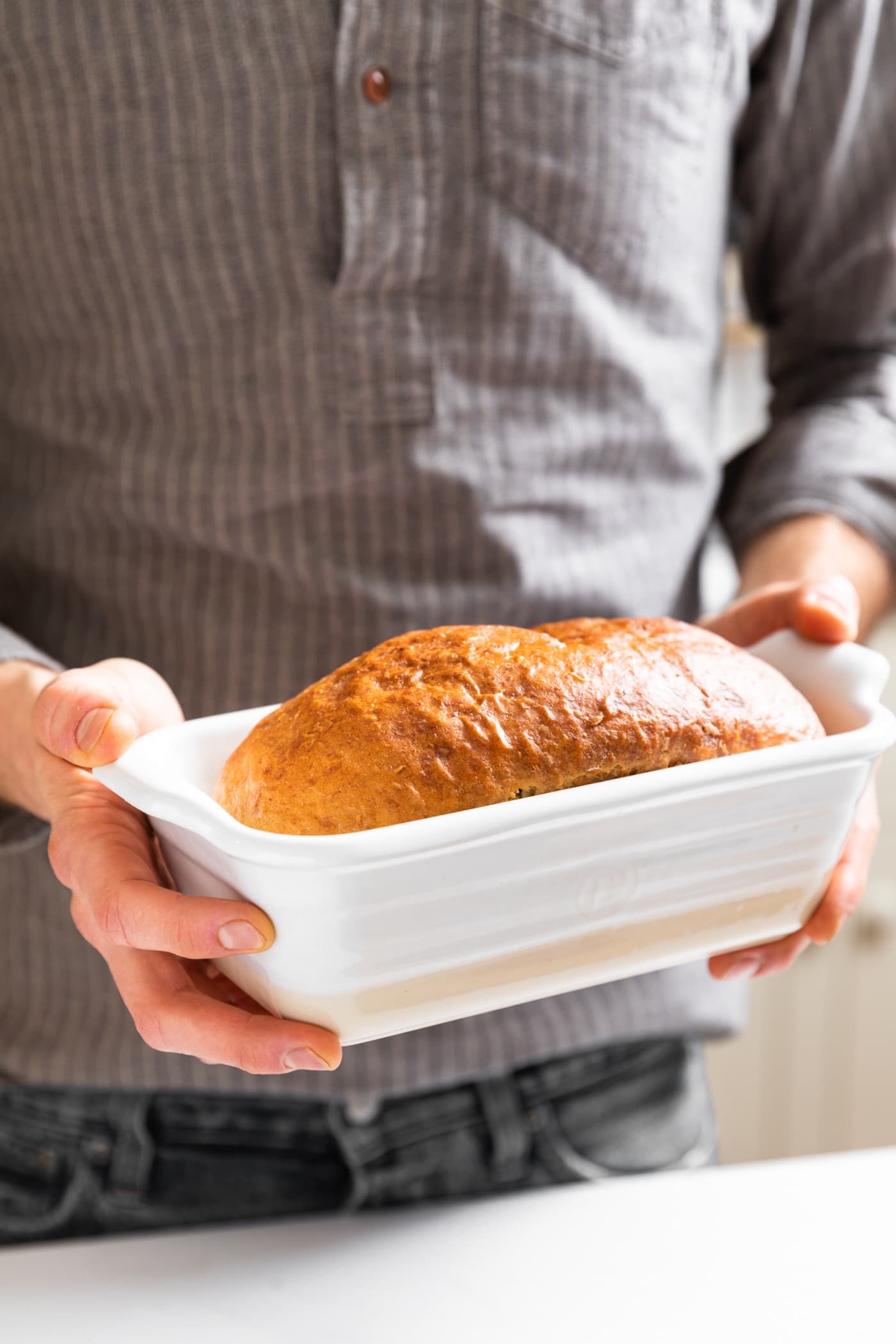 Loaf of golden colored bread sitting in white loaf pan being held by person with gray shirt on