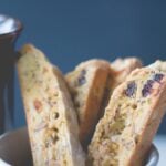 White bowl filled with slices of fruit and nut biscotti dotted with pieces of dried fruit with coffee cup in background