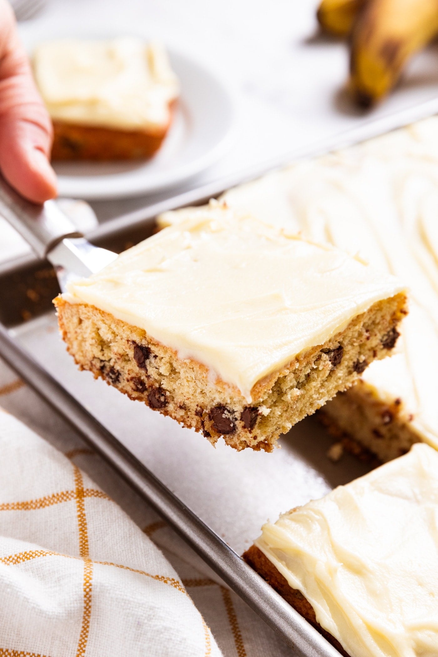 Hand holding spatula holding up a square of chocolate chip banana bar with rest of banana bars still in pan below with bananas to the side.