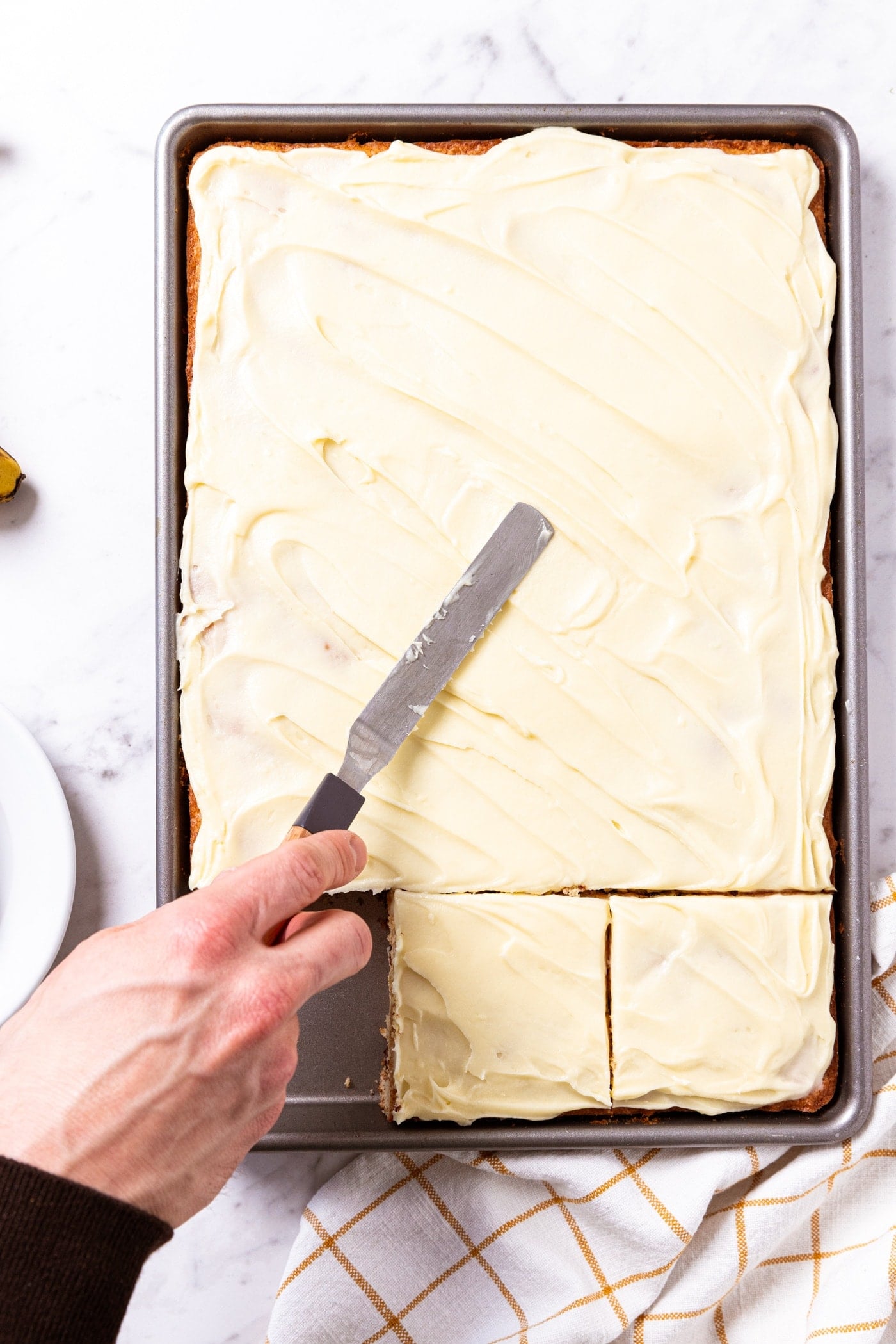Hand using offset spatula to spread cream cheese frosting over the top of pan of chocolate chip banana bars sitting on white marble surface.