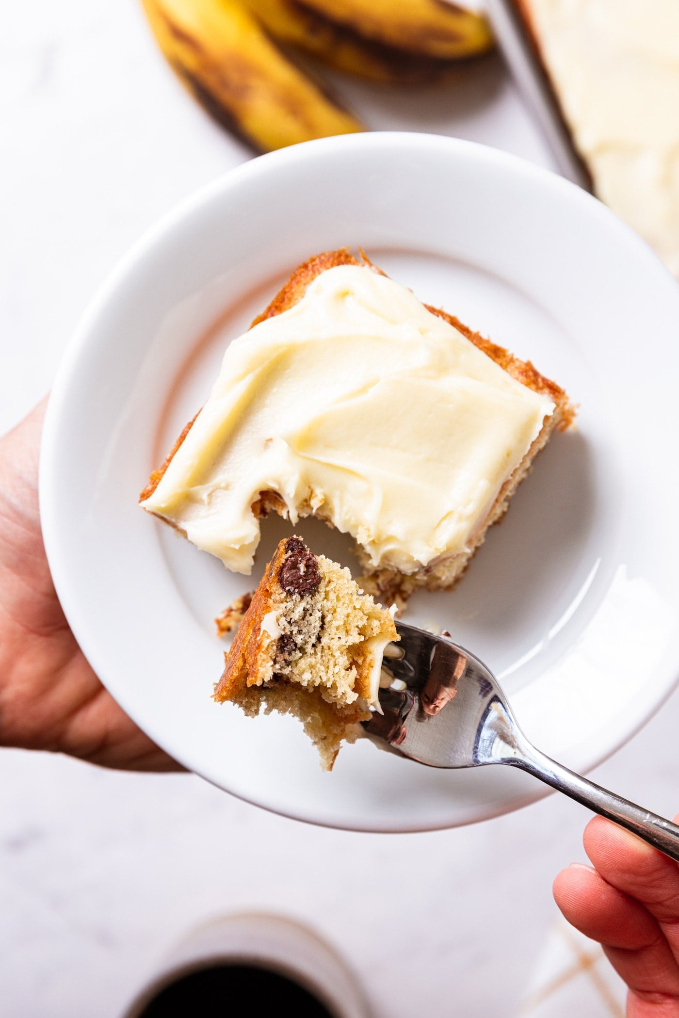 Hand holding fork with bite-full of chocolate chip banana bar with piece of banana bar on white plate below.