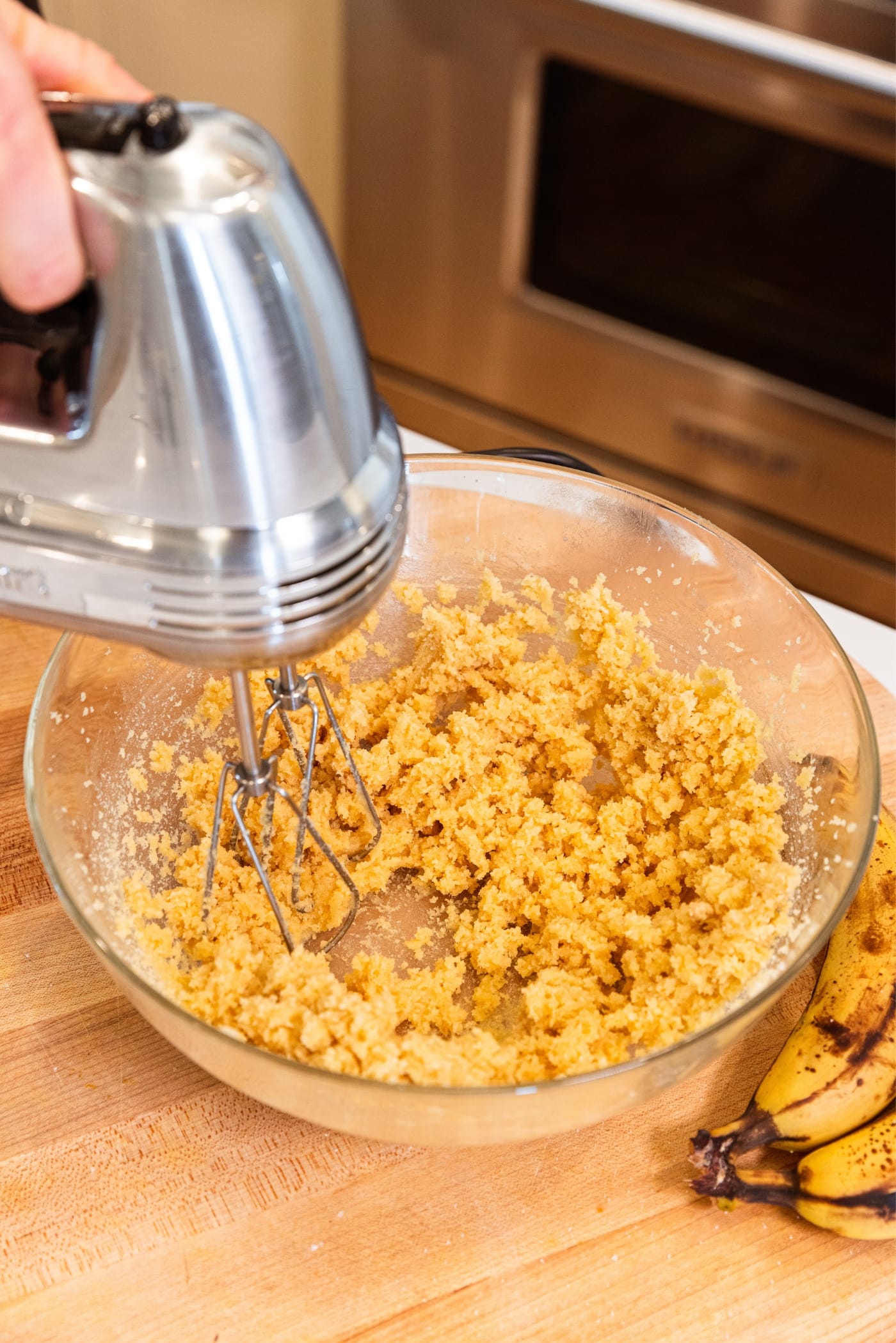 Hand mixer mixing together butter and sugar mixture on wood cutting board with unpeeled bananas in background.