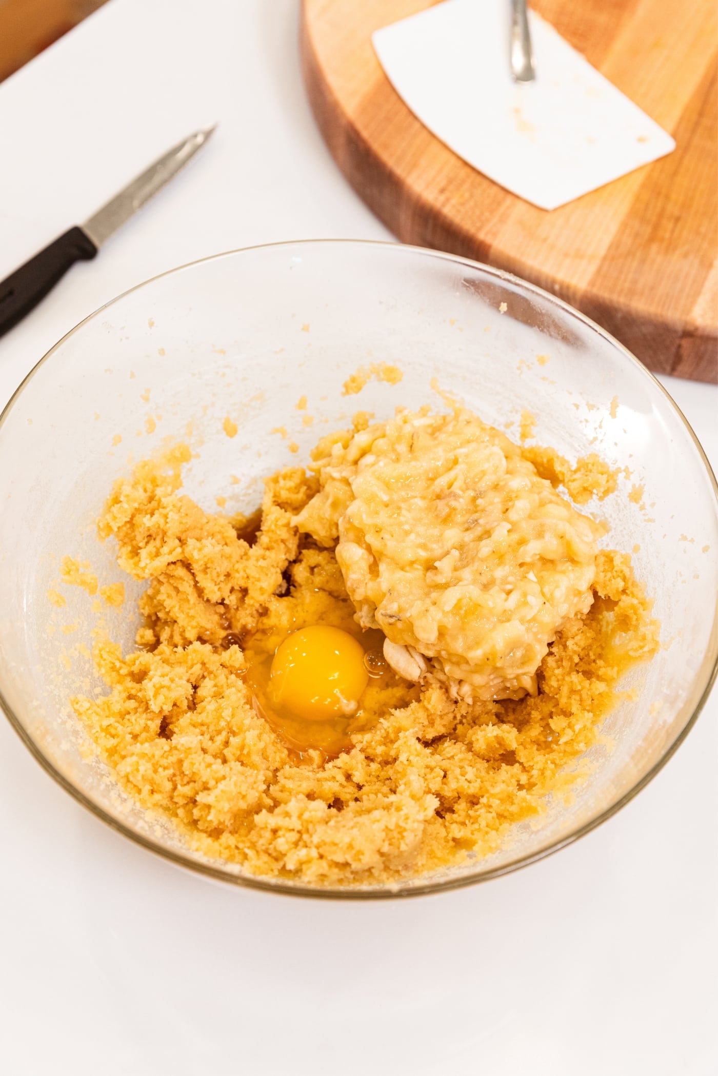 Glass bowl with mixed sugar and butter mixture underneath with raw egg cracked into bowl along with mashed banana.