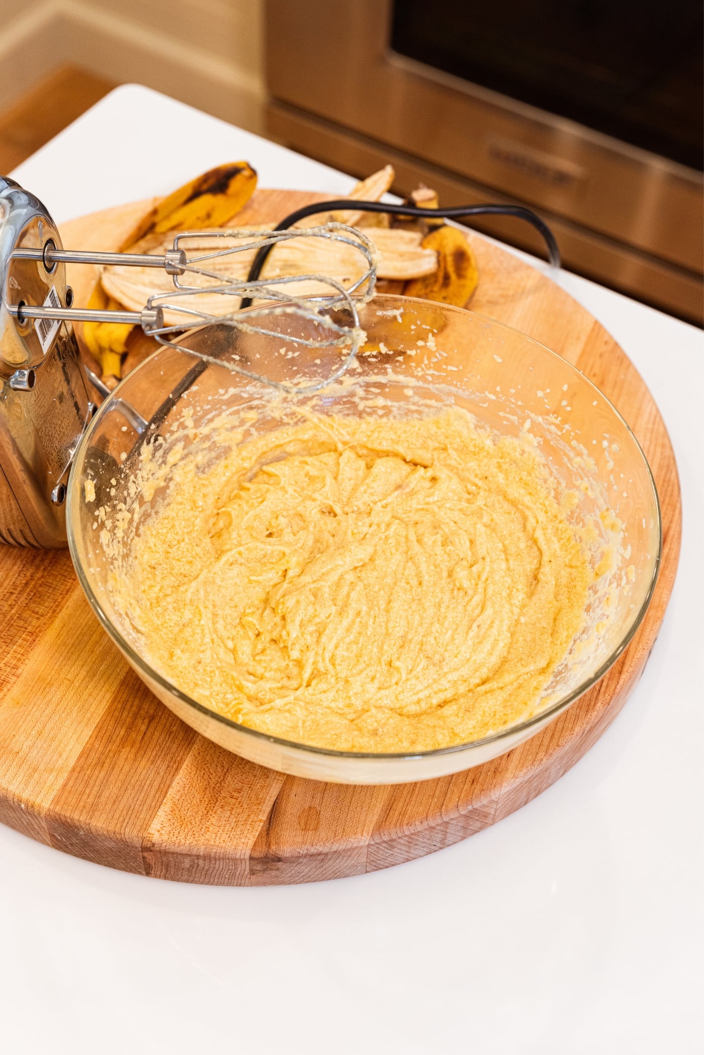 Glass bowl with mixed banana bars batter inside with hand mixer resting on wood cutting board with banana peels in background.