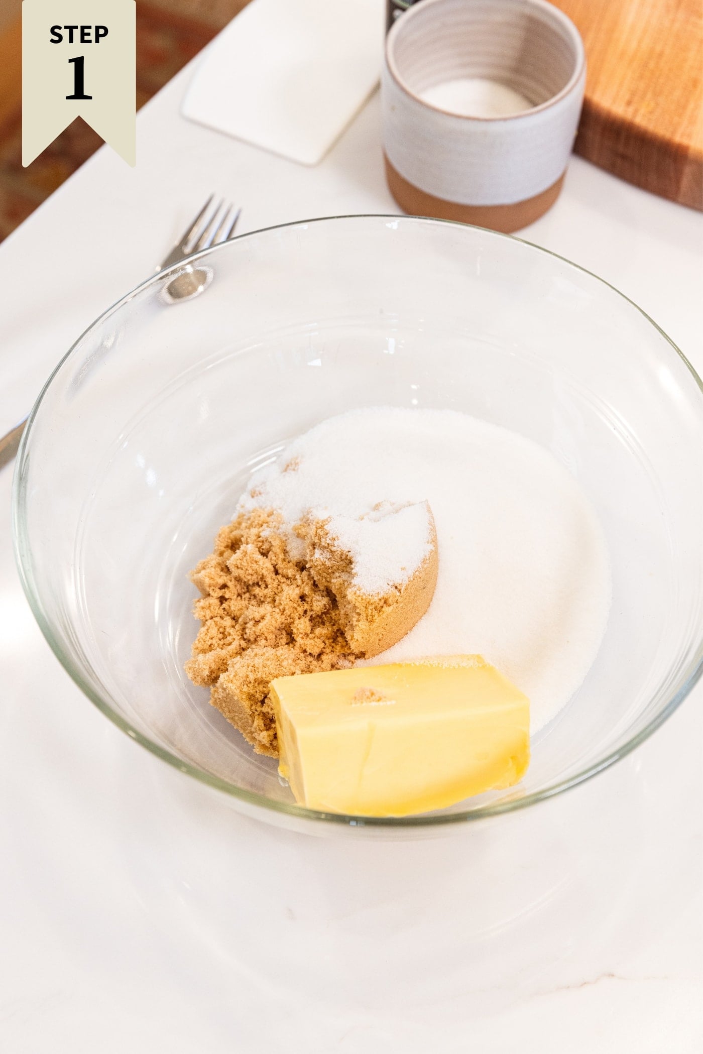 Glass bowl sitting on white surface filled with white sugar, brown sugar, and butter before being mixed together with hand mixer.