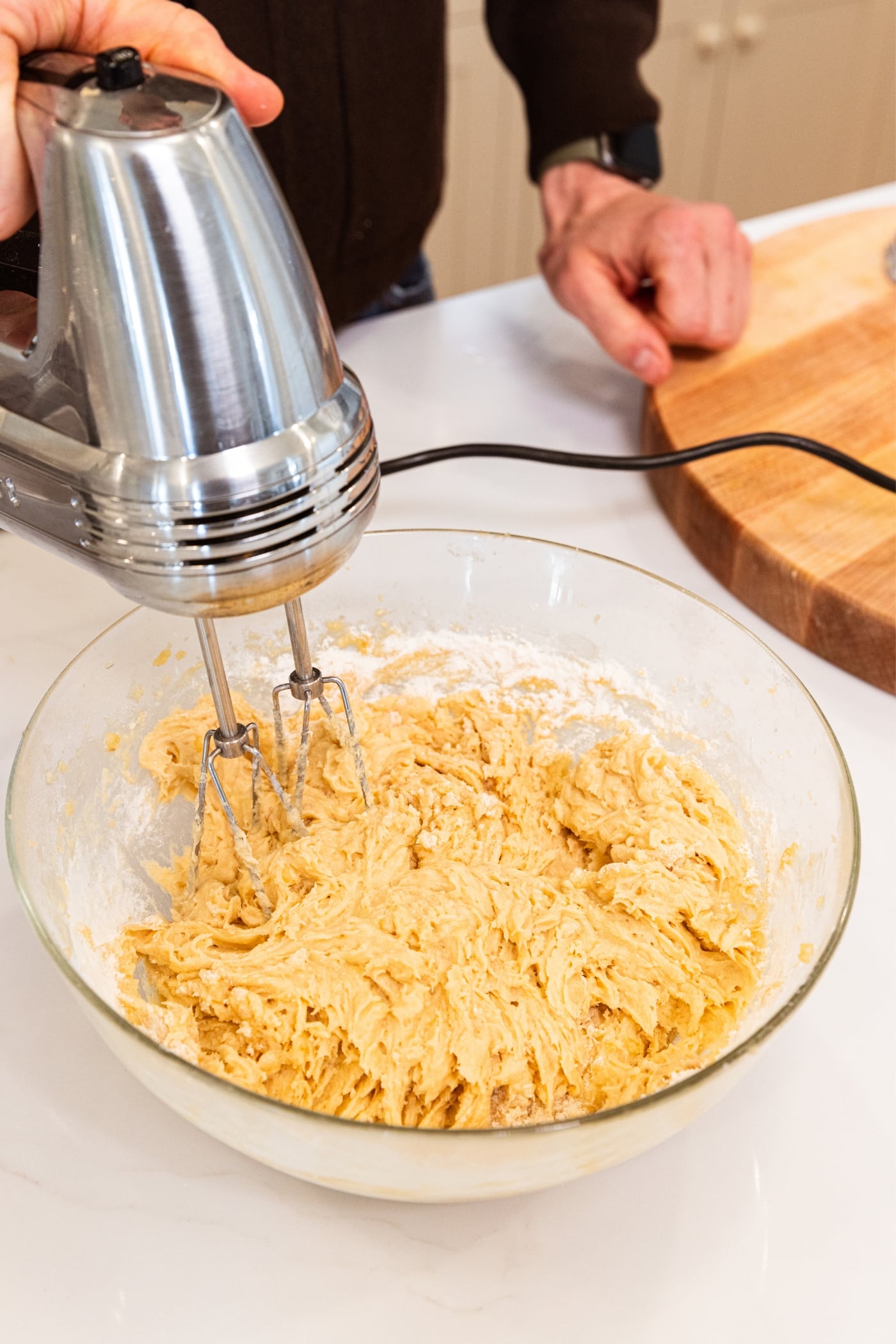 Hand mixer mixing flour into wet ingredients in a glass bowl creating a wet batter with hand resting on wood cutting board in background.
