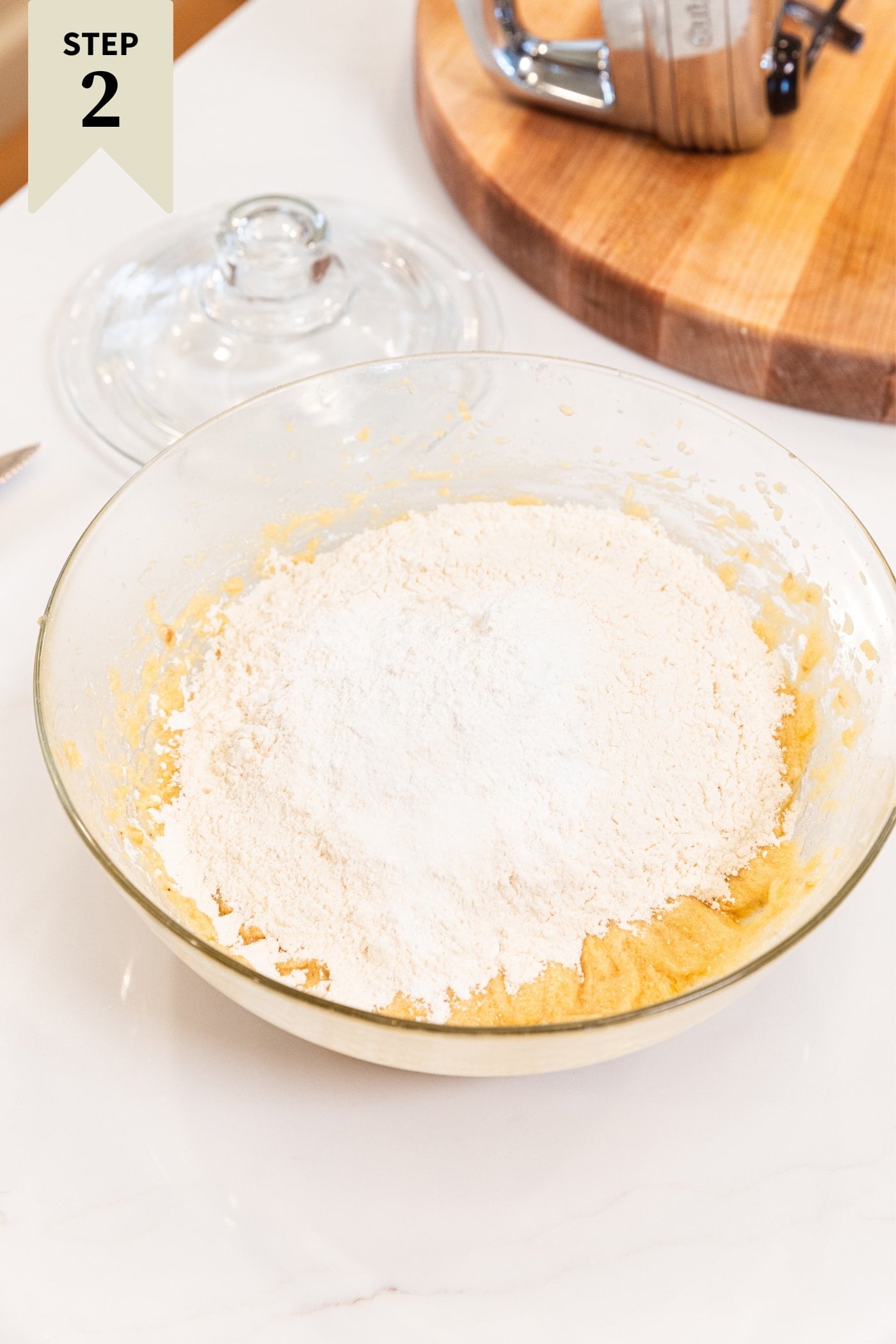 Glass bowl sitting on white surface filled with mixed wet ingredients with flour on top right before being mixed together with hand mixer.