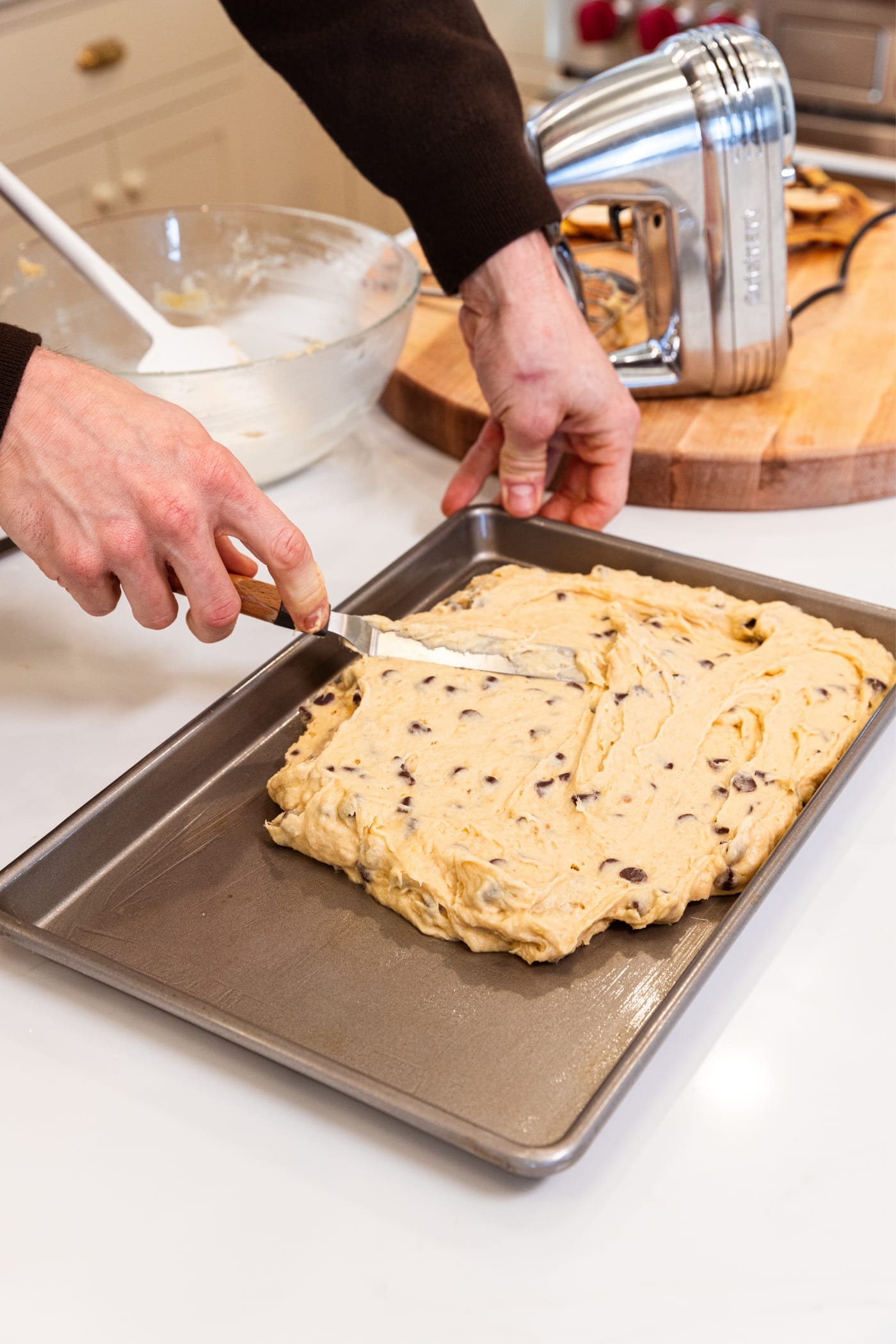 Hand using offset spatula to spread banana bar batter dotted with chocolate chips into metal baking pan on white surface.