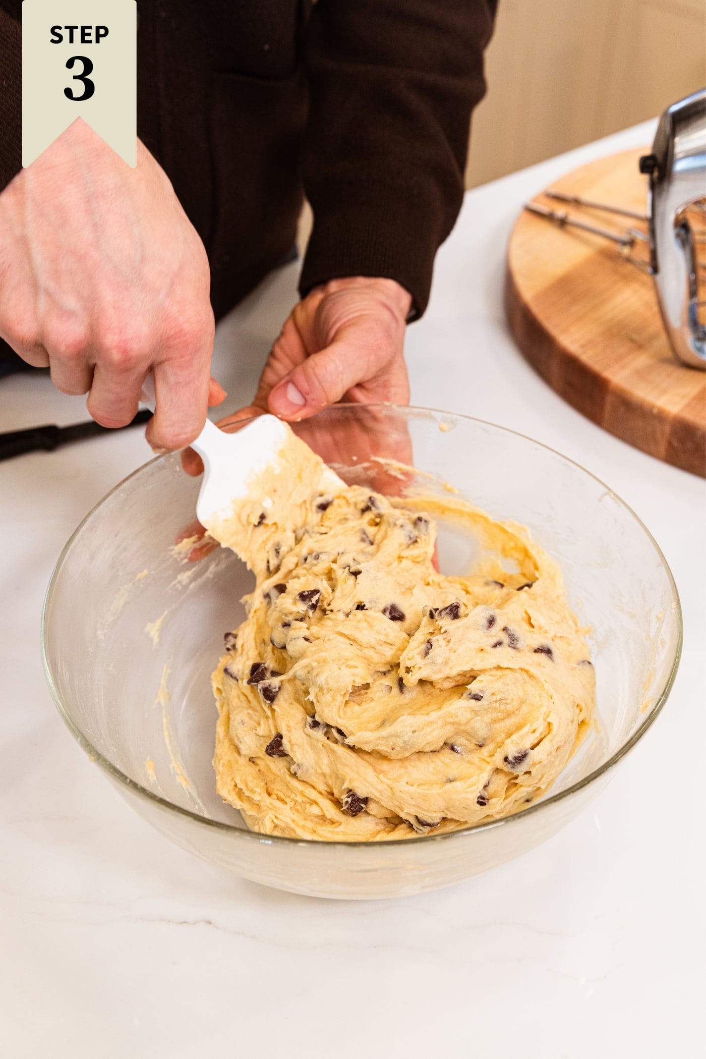 Hand using white spatula to mix together chocolate chips with light brown colored batter.