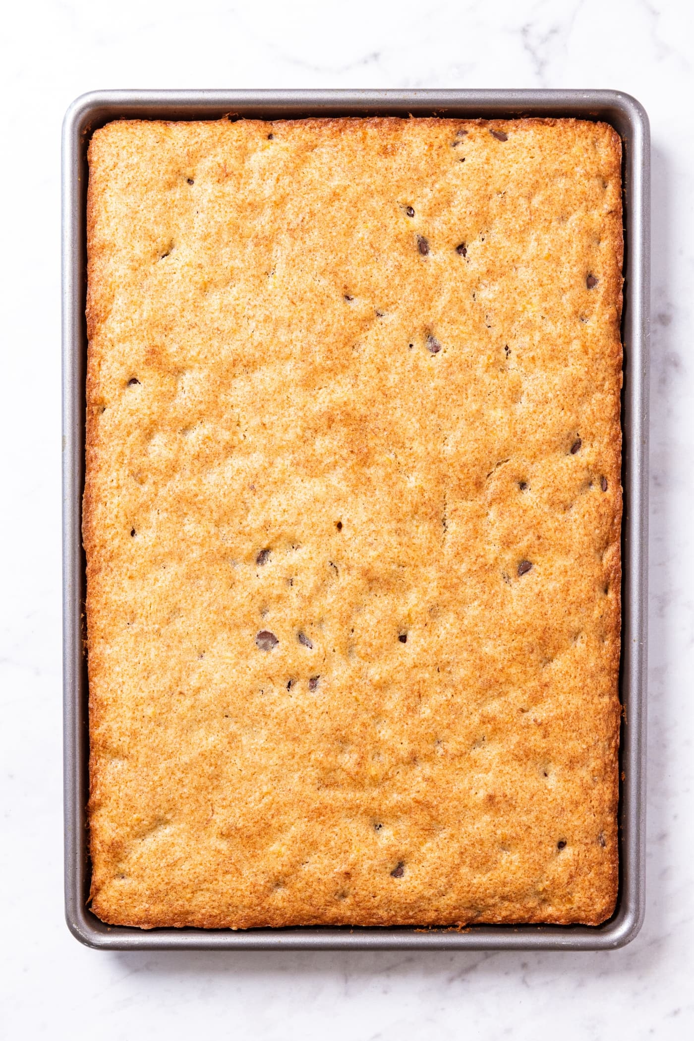 White marble surface with metal baking dish with baked banana bars in the pan showing golden top and slightly pulling away from the edges of the pan.
