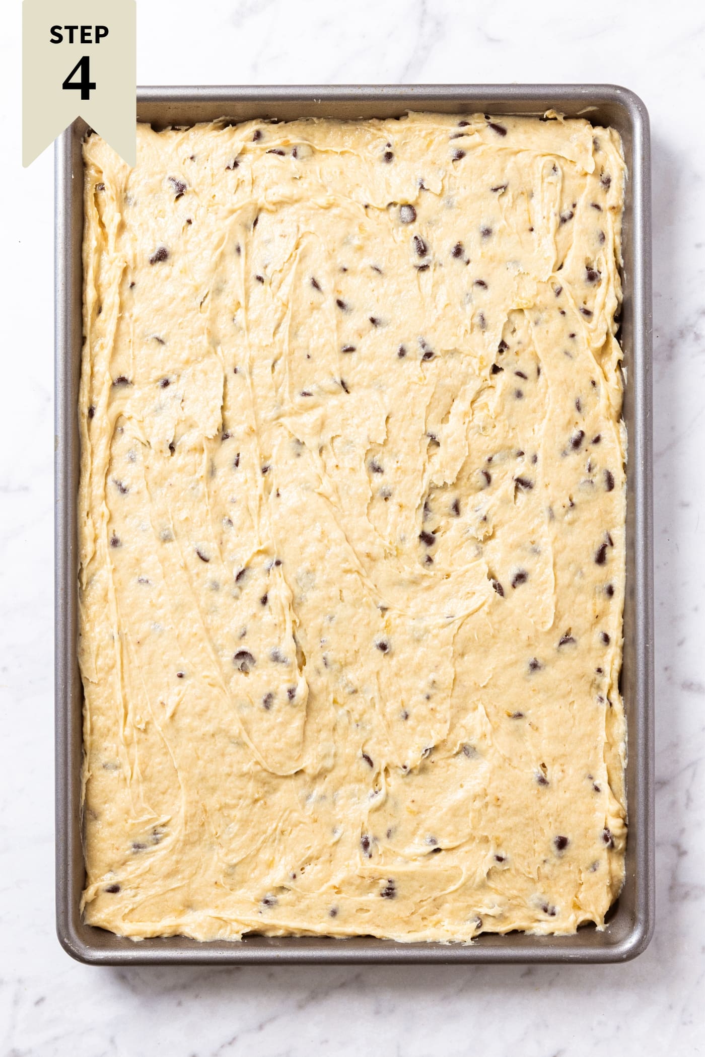 White marble surface with metal baking pan filled with unbaked banana bars dotted with chocolate chips throughout right before going in the oven.