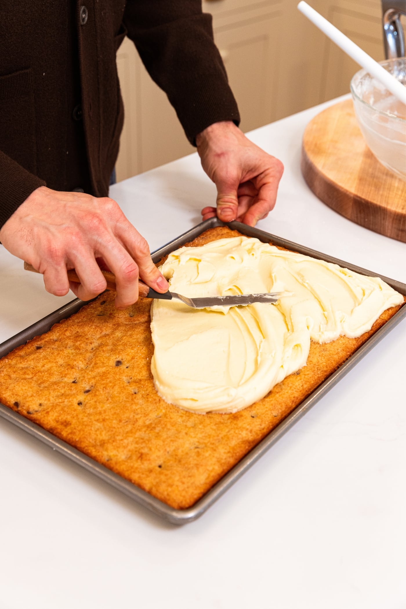 Hand using offset spatula to spread out cream cheese frosting on top of cooled baked banana bars on white surface.