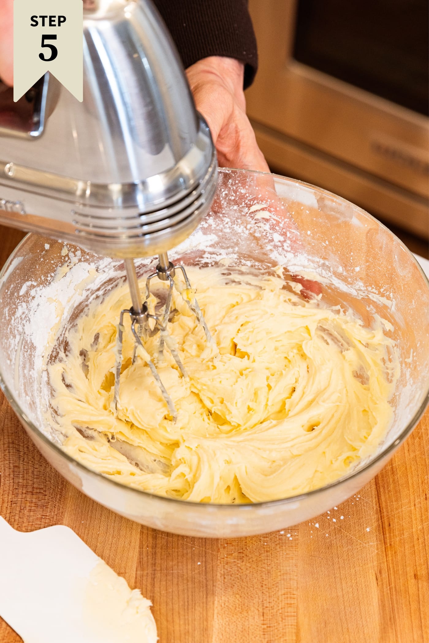 Hand mixer mixing together ingredients needed for cream cheese frosting with light creamy texture on wood cutting board.