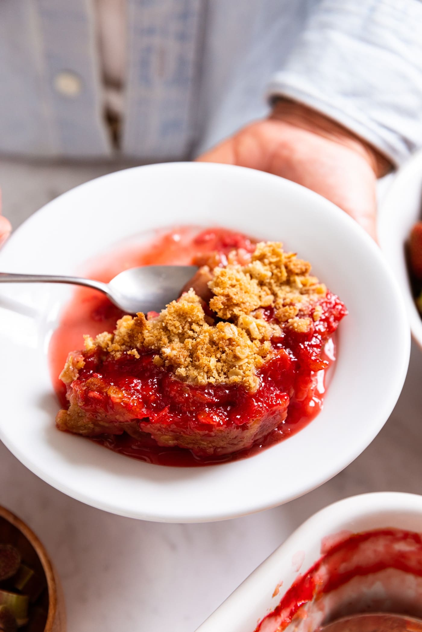 Hands holding small white plate with serving of strawberry rhubarb crumble with spoon scooping a piece.
