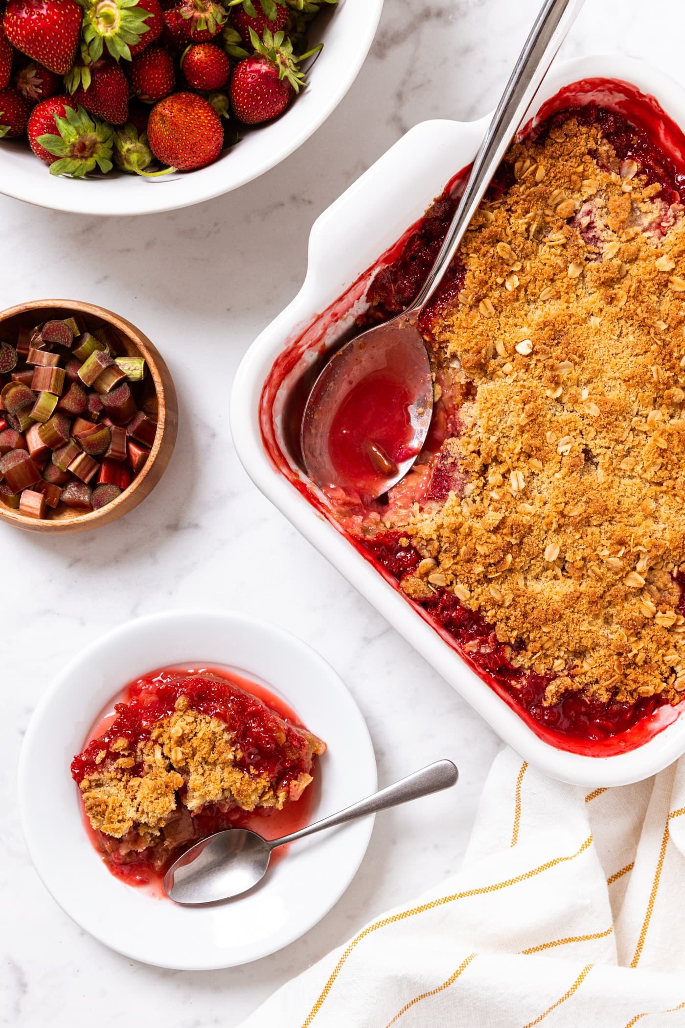 White marble surface with baking dish filled with strawberry rhubarb crumble with spoon taking a serving out and one serving sitting on a white plate with chopped rhubarb and strawberries to the sides.