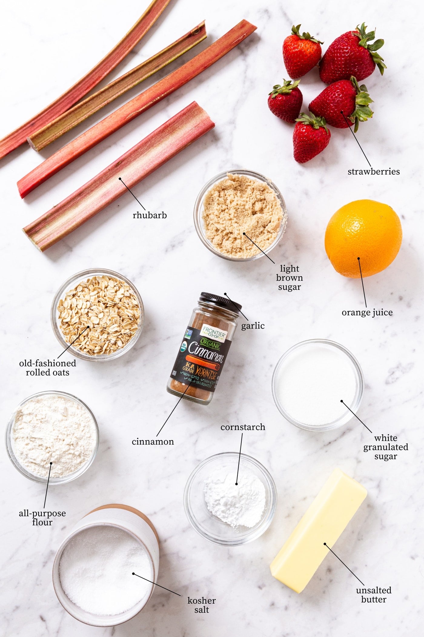 White marble surface with all ingredients needed to prepare strawberry rhubarb crumble including raw rhubarb, strawberries, orange, sugar, oats, cinnamon, and more.