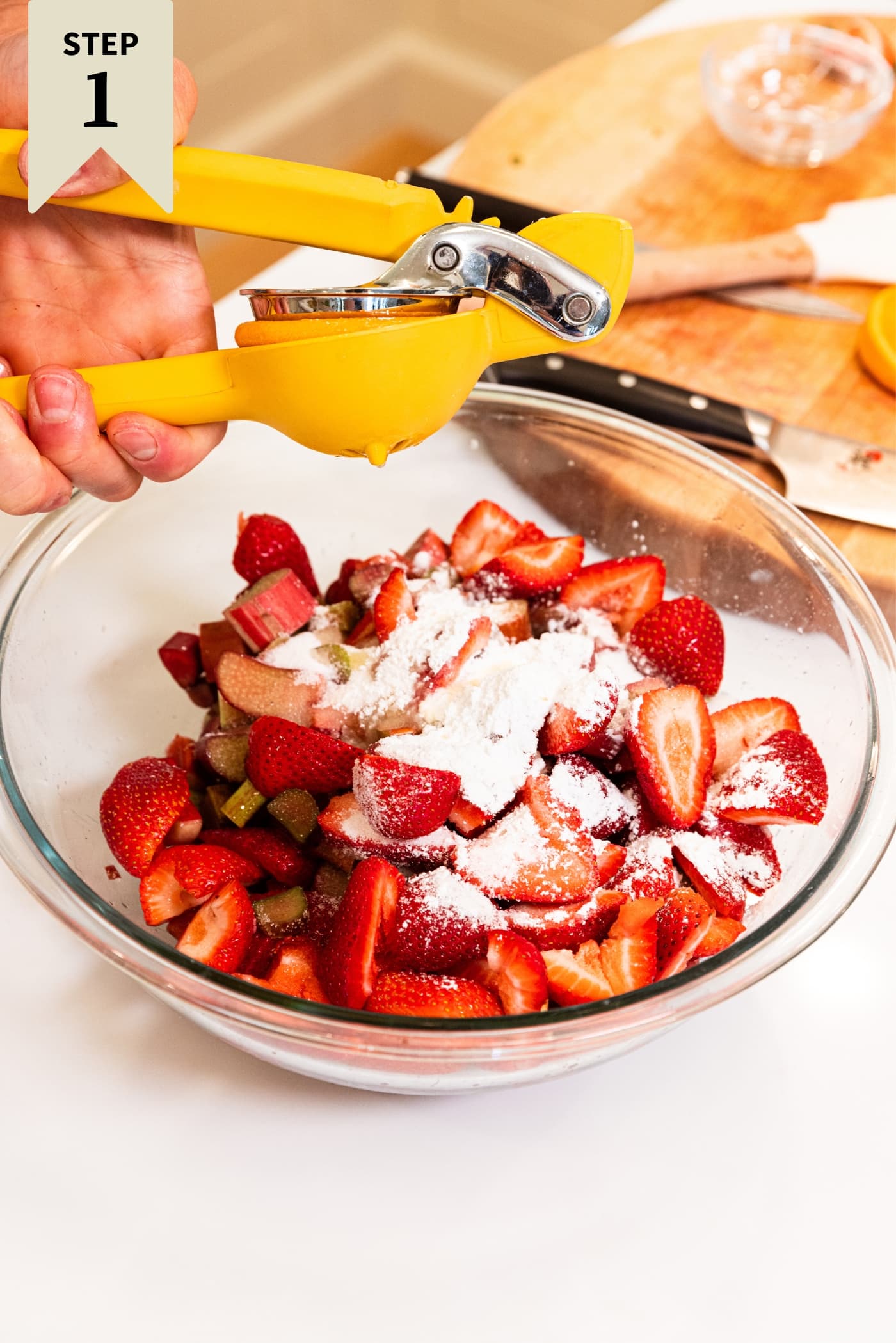 Hand holding lemon juicer squeezing lemon juice on top of mixture of chopped rhubarb and strawberries with cornstarch sprinkled on top all in glass bowl on white surface.