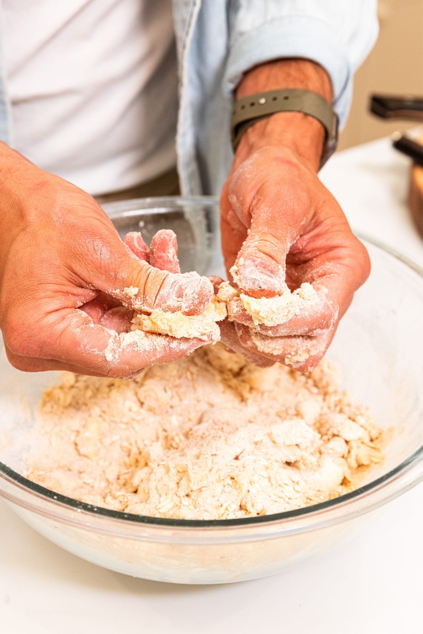 Two hands pressing together butter with oat and flour mixture to smash the butter into small pieces into large glass bowl.