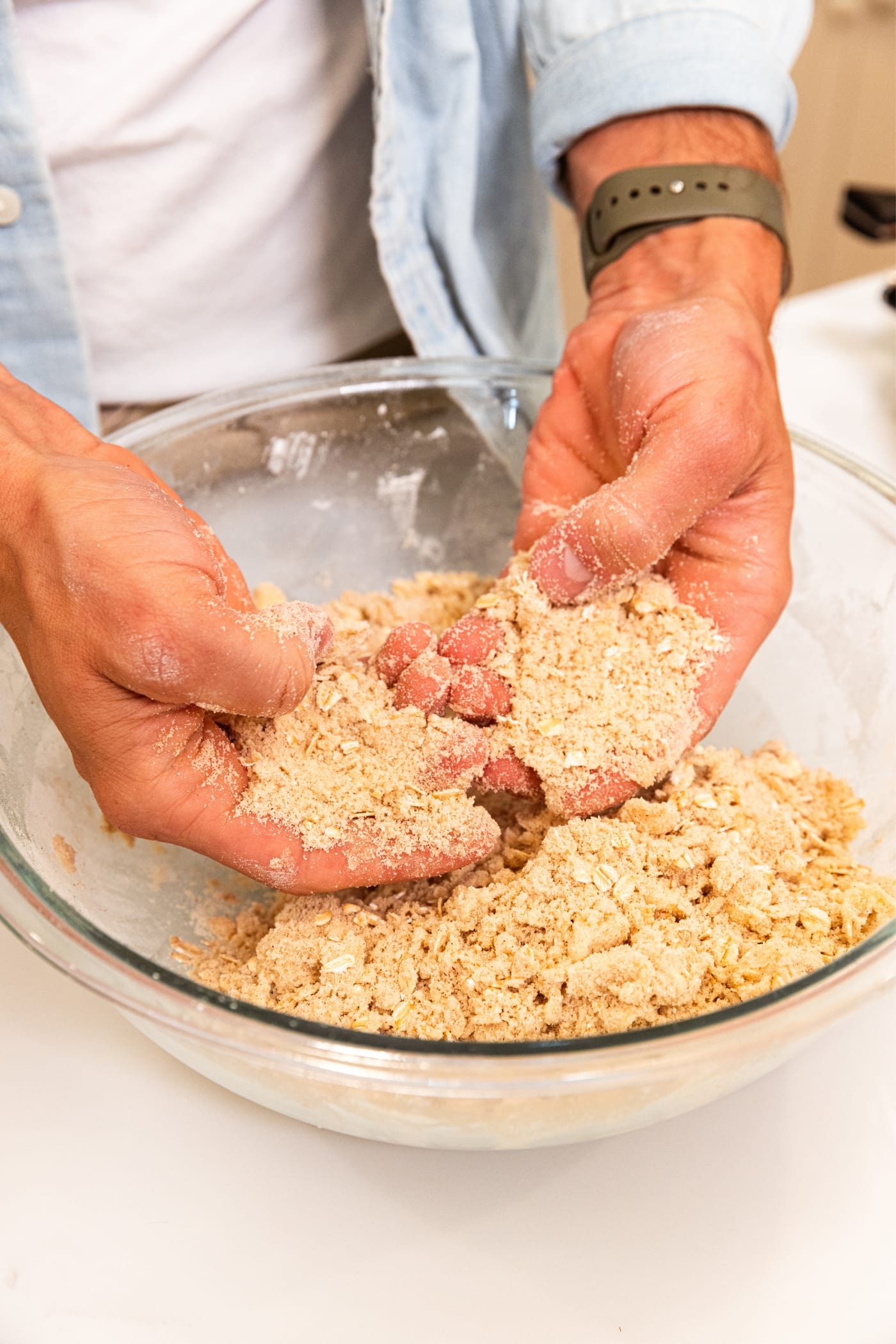 Two hands showing sandy texture of crumble topping in glass bowl on white surface.